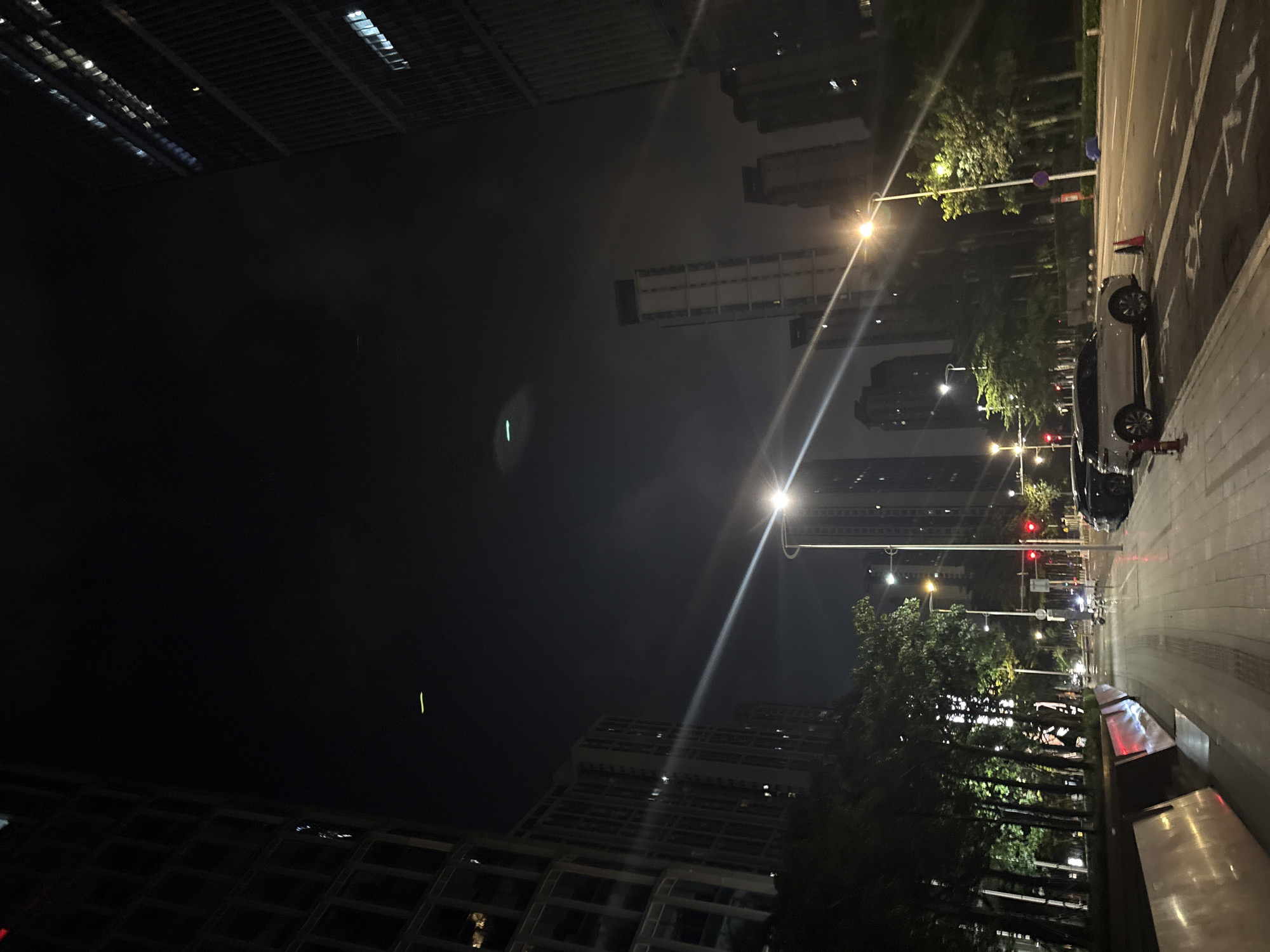 This is a nighttime, slightly low-angle shot taken in an urban setting, possibly Haizhu District, China. The image is dominated by a dark, misty sky, with several tall buildings visible in the background. Streetlights cast a warm glow on the scene, illuminating a paved walkway and a road.
On the right side of the image, a white car is parked, and a red traffic cone is placed on the road. The car's headlights are on, suggesting it might be ready to move or has recently stopped. Further down the road, a traffic light is visible, showing a red signal, and a few other vehicles are present. Trees line the walkway, their leaves catching the light from the streetlamps, creating a dappled effect.
The overall atmosphere is quiet and serene, with the artificial lights piercing the darkness. The mist in the air softens the outlines of the buildings and creates a sense of depth. No people are clearly visible in the image, contributing to the tranquil mood. The time of day is clearly night due to the artificial lighting and the dark sky. The presence of streetlights, tall buildings, and a paved road strongly indicates an urban environment.