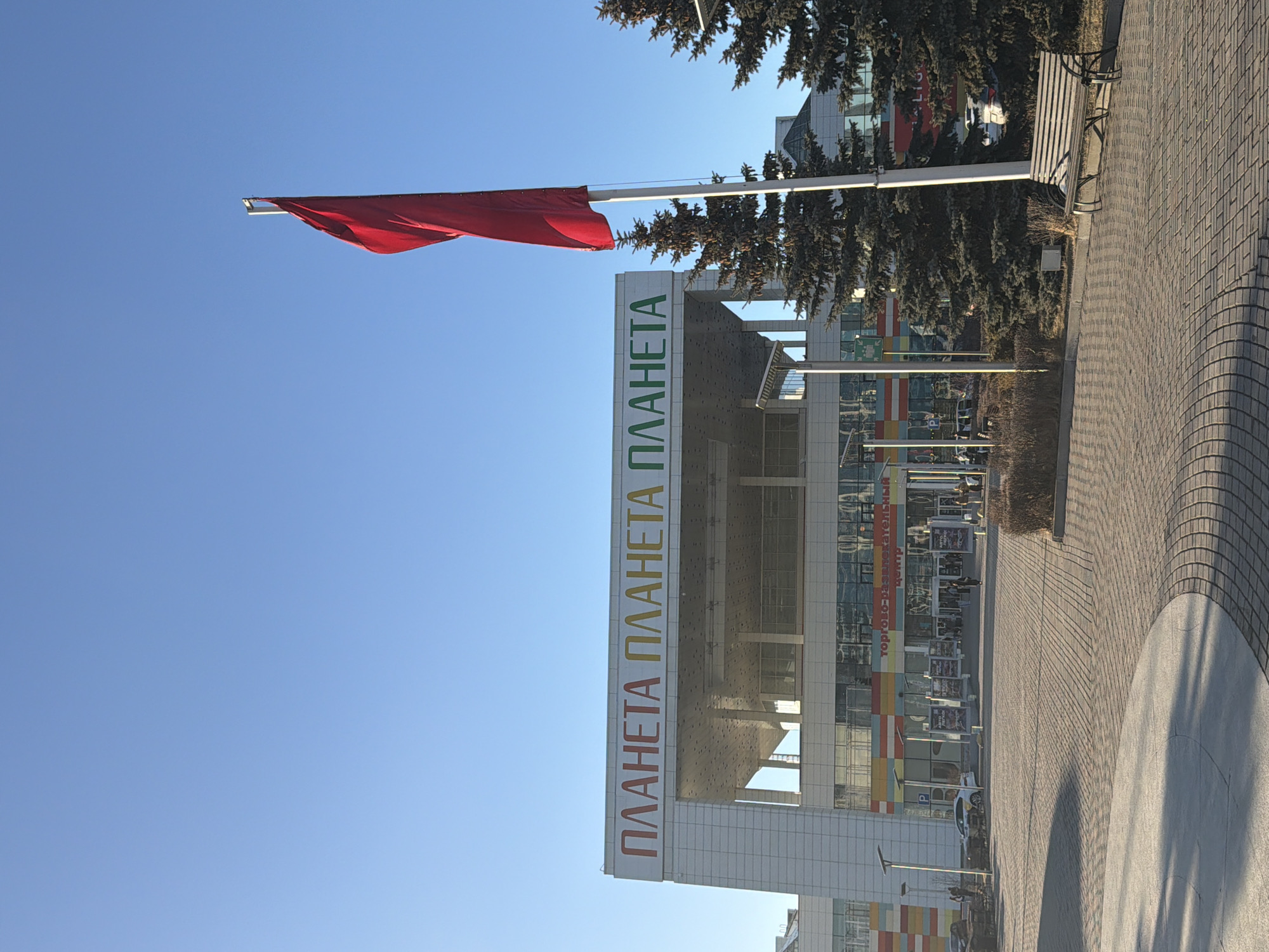 The image shows a red flag on a white pole with a building behind it in Krasnoyarsk, Russia. The building has the word "ПЛАНЕТА" written on it vertically in two colors. There is a sidewalk in the foreground with a bench beside the building. The sky is blue.