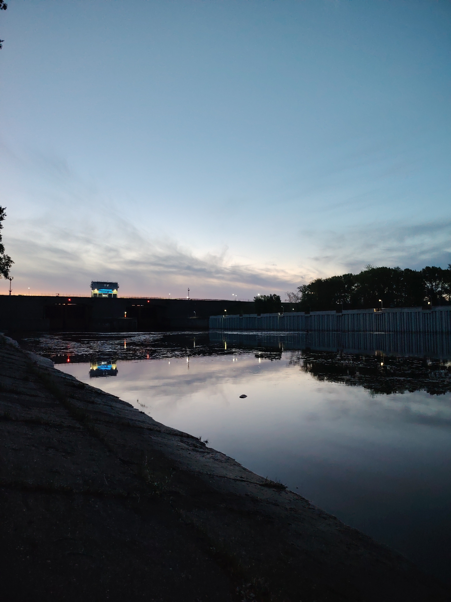 The image depicts a body of water, possibly a river or lake, in the city of Chaikovsky, Russia. A dam or barrier with a building and support structures spans the water's width in the distance. The building is illuminated with blue lights. The sky is dusky, indicating either dawn or dusk. The water reflects the sky and the building's lights. The foreground consists of a concrete embankment along the water's edge. The lighting is low, suggesting nighttime or twilight conditions. Trees line the far bank of the water.