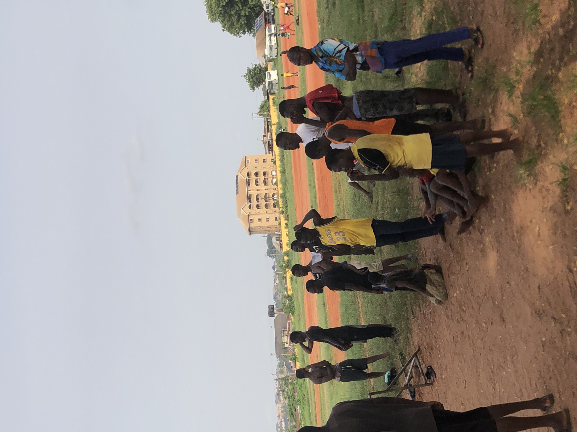 This image captures an outdoor scene in Ukwu-Abwa, Nigeria, under a bright, clear sky, suggesting daytime, possibly morning or late afternoon. The setting is an expansive open field, characterized by sections of reddish-brown dirt juxtaposed with patches of green grass, indicative of a sports ground or recreational area.
The main subjects are a group of approximately 10-15 young males, appearing to be teenagers or young adults, all of whom are Black. They are distributed across the midground and foreground, standing in various casual poses. Some appear attentive, possibly listening or observing, while others are more relaxed. Their attire is casual; most wear t-shirts and shorts, though one individual sports a blue patterned shirt with blue trousers, another has a red cloth draped over their shoulder, and a prominent figure wears a yellow basketball jersey with "LAKERS 23" clearly visible. Several individuals are barefoot or wearing sandals. More figures are visible in the distance, closer to the background structures.
In the background, there is a multi-story building with a light brown facade, along with other smaller buildings and what appear to be white vans or buses. Lush green trees are also scattered across the horizon, with a large tree dominating the upper right portion of the frame. On the dirt ground, a small, rudimentary wooden structure is visible near the bottom center, alongside a small, dark object that could be a bird.
The scene depicts a gathering, possibly for a sports practice, a training session, or a community event. The group's attentive yet relaxed demeanor suggests they might be receiving instructions or preparing for an activity. The "LAKERS 23" jersey indicates an interest in sports, although the specific nature of the activity on the dirt field is not definitively shown. The overall mood appears calm and focused.
