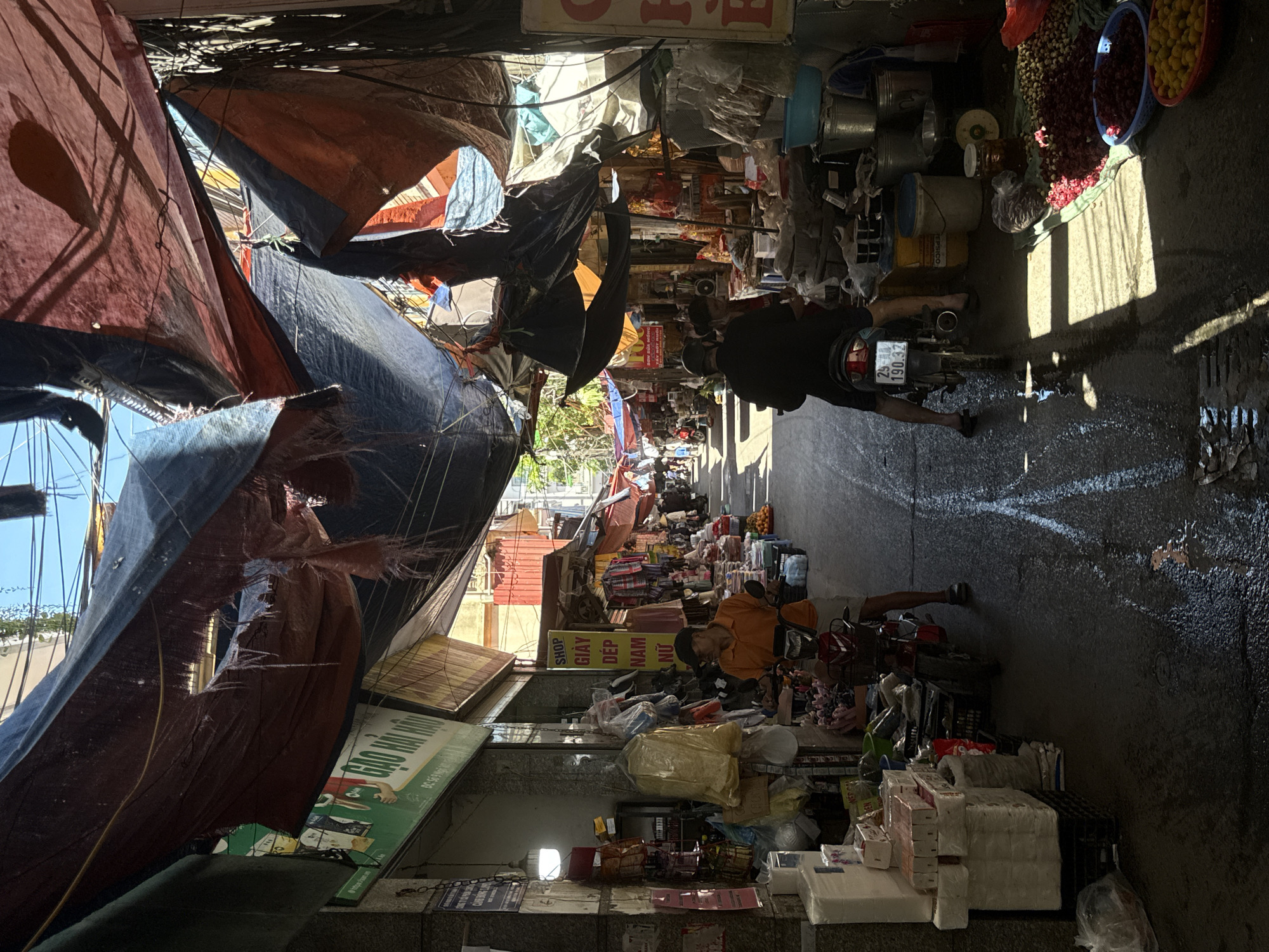 The image shows a crowded street scene in Hanoi, Vietnam, likely a market area. The street is narrow and lined with vendors selling various goods. Awnings and tarps are stretched overhead, providing shade. People are moving about, including one individual on a motorbike. Goods for sale include produce, household items, and footwear. The street surface is wet, reflecting sunlight.