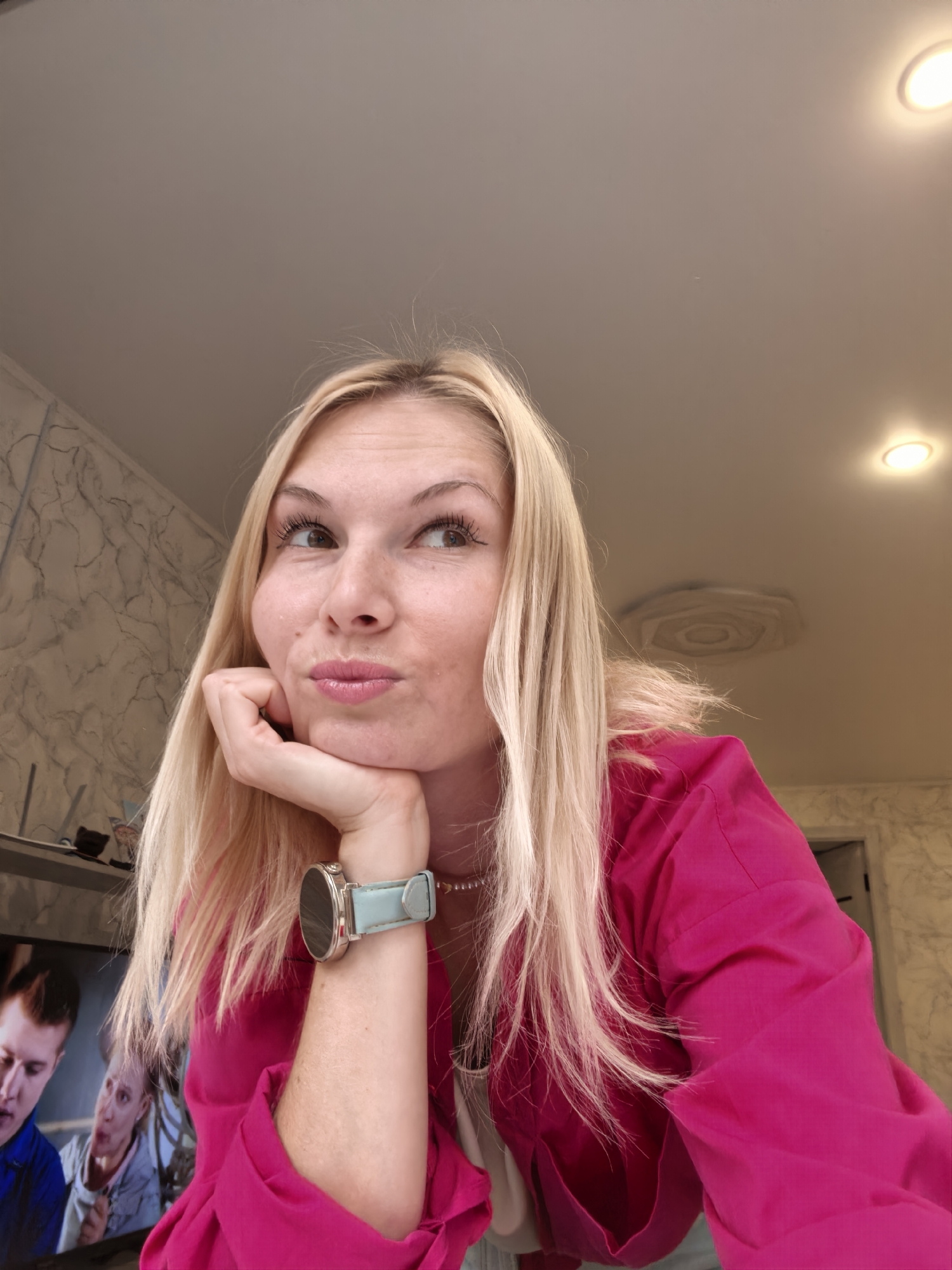 A blonde woman with her chin resting on her hand is looking to the right. She is wearing a bright pink jacket and a light blue watch. In the background, a television screen displays two men, and the wall has a marbled pattern. The ceiling has recessed lighting fixtures. The scene appears to be indoors, possibly in a residential setting. There is no information in the image to suggest it is located in Sterlitamak, Russia.