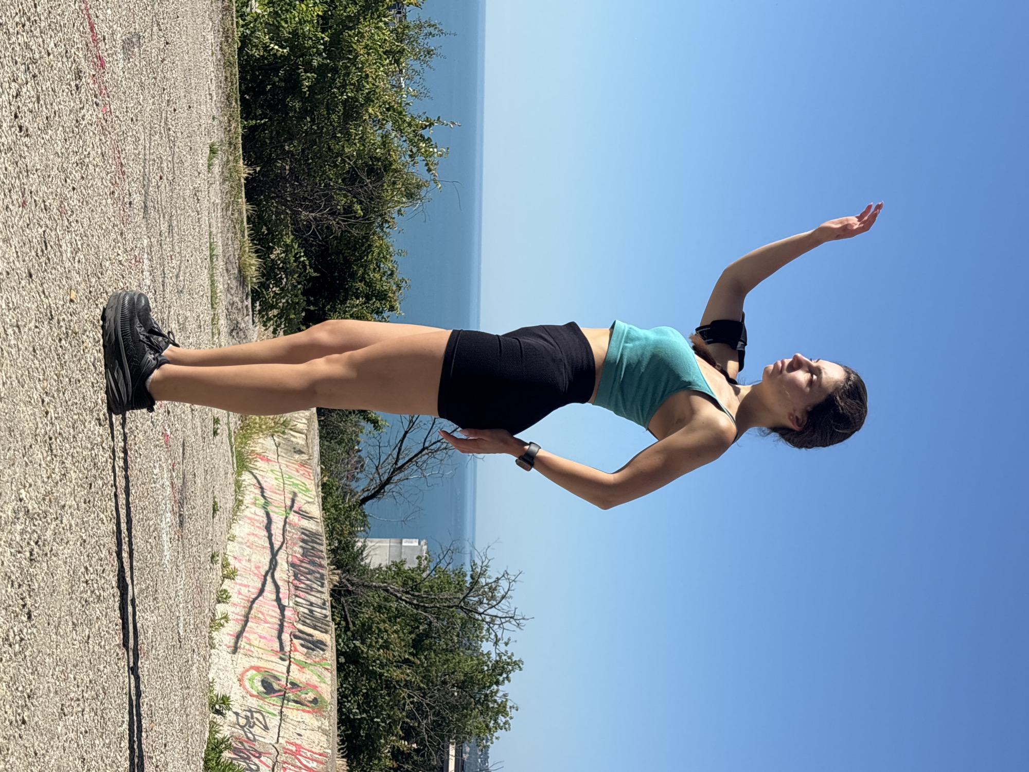 In Varna, Bulgaria, a young woman stands on a concrete surface with a view of the sea. She is wearing a teal crop top, black shorts, and black sneakers. On her left arm, she has a black armband with a device inside, and a smartwatch is visible on her right wrist. Her right hand is extended upwards, and her left arm is bent behind her back. Behind her, green bushes and trees lead down to the blue sea, which stretches to the horizon under a clear blue sky. A concrete wall with graffiti is visible in the foreground.