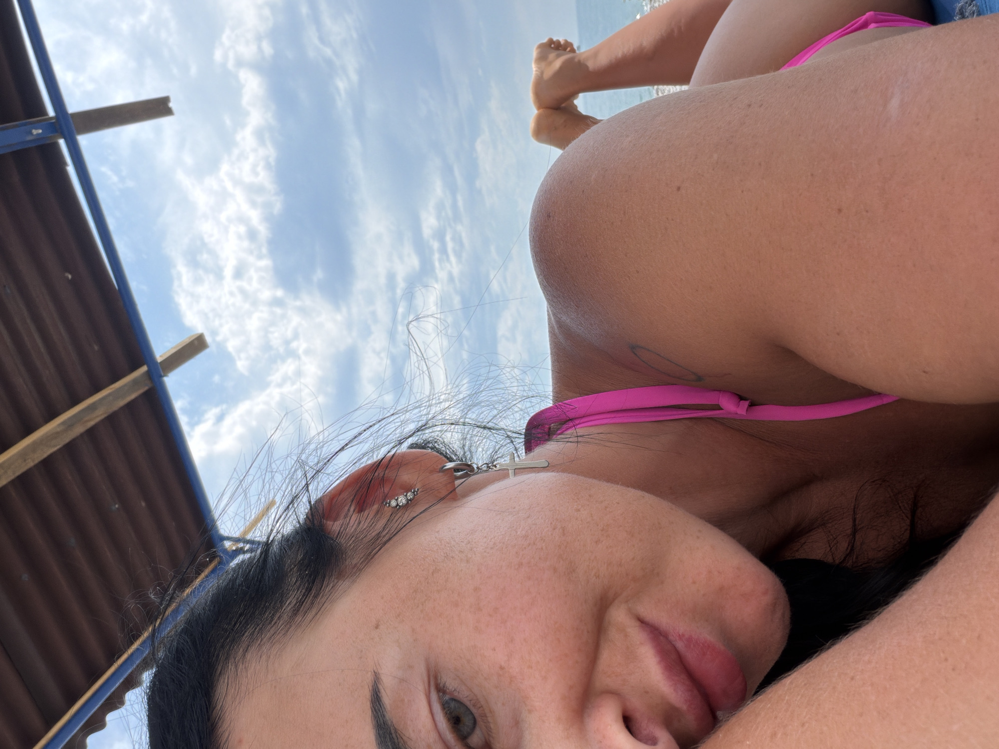 This is a close-up, low-angle, outdoor photograph taken from a first-person perspective. The scene appears to be a beach or seaside location, possibly Sochi, Russia, given the context.
The primary subject is a person, with the camera positioned close to their face. We see a portion of their face, including an eye, lips, and cheek with freckles. They are wearing a pink bikini top, and a silver earring with small gems and a cross charm is visible on their ear. Dark hair is also visible.
In the background, the sky is visible with scattered clouds. There are also elements of what appears to be a beach structure, possibly a sunshade or shelter, with corrugated roofing and wooden beams. The person is lying down, with their legs and feet visible in the upper portion of the image, suggesting they are relaxing or sunbathing. The water is visible in the distance.
The time of day appears to be daytime, with bright natural light illuminating the scene. The weather seems pleasant, with a clear sky and clouds. The overall mood conveyed is relaxed and summery. There is no visible text in the image.