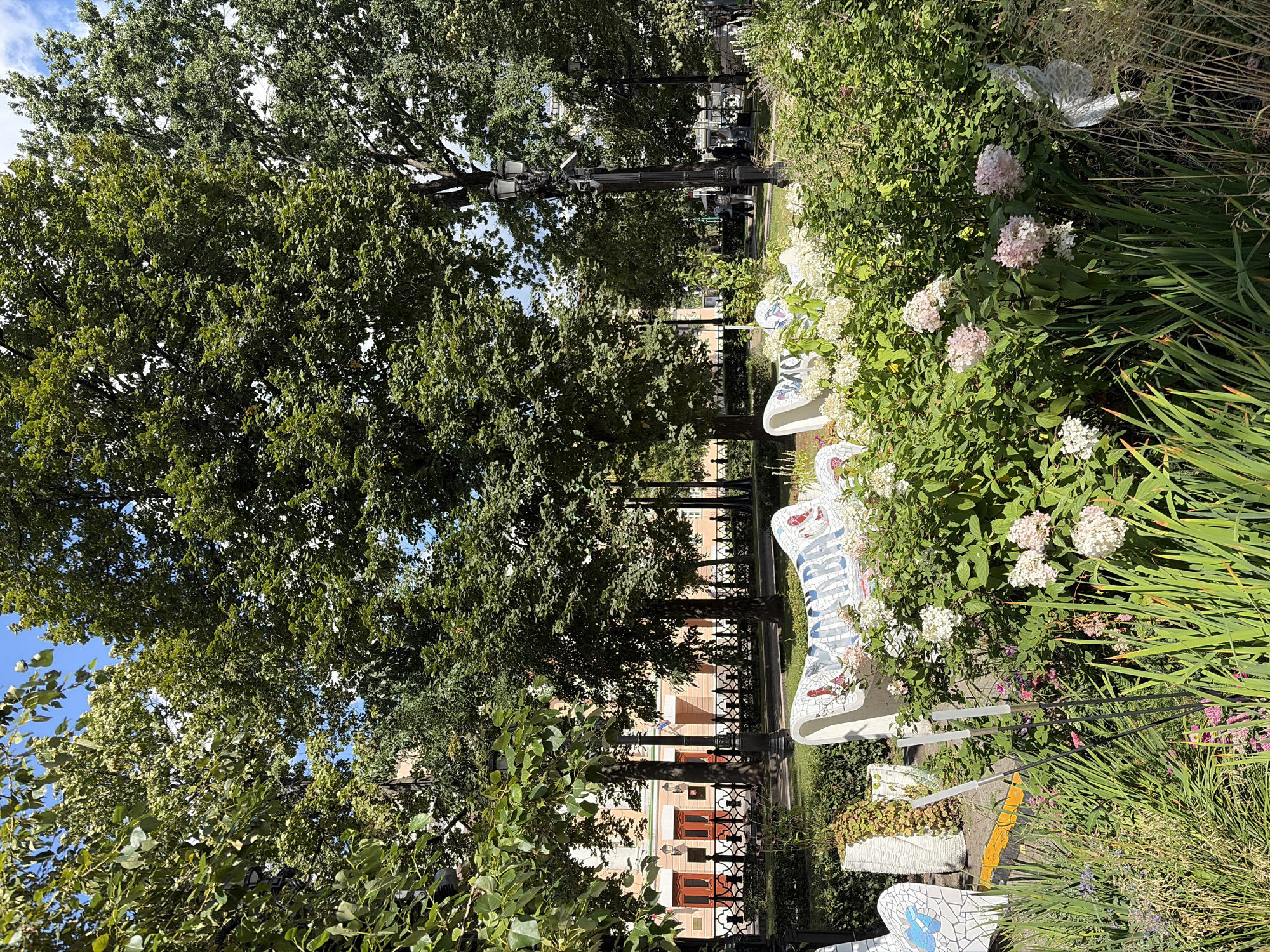 The image captures a sunny day in a park, likely Moscow, Russia, given the architectural style visible in the background. In the foreground, a white mosaic bench is decorated with colorful tiles, displaying text. Adjacent to the bench is a lush green garden filled with various plants and flowers, including white hydrangeas. Beyond the garden, a black ornate fence separates the park from a street lined with trees. In the background, a building with a light-colored facade and red doors is visible. The scene is serene and depicts a tranquil urban park setting.