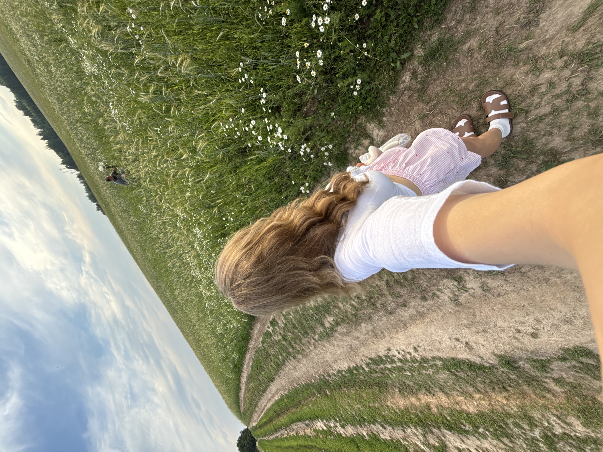 A person with long, wavy blonde hair is taking a selfie while walking on a dirt path through a field of green grass and white wildflowers. The person is wearing a white crop top and a pink and white striped skirt, with white socks and brown sandals. Another person is visible in the distance in the field, appearing to be picking flowers. The scene is set in a rural area with a gently sloping hill and a line of trees in the background. The sky is blue with scattered white clouds. The location is identified as Сновицы, Russia.