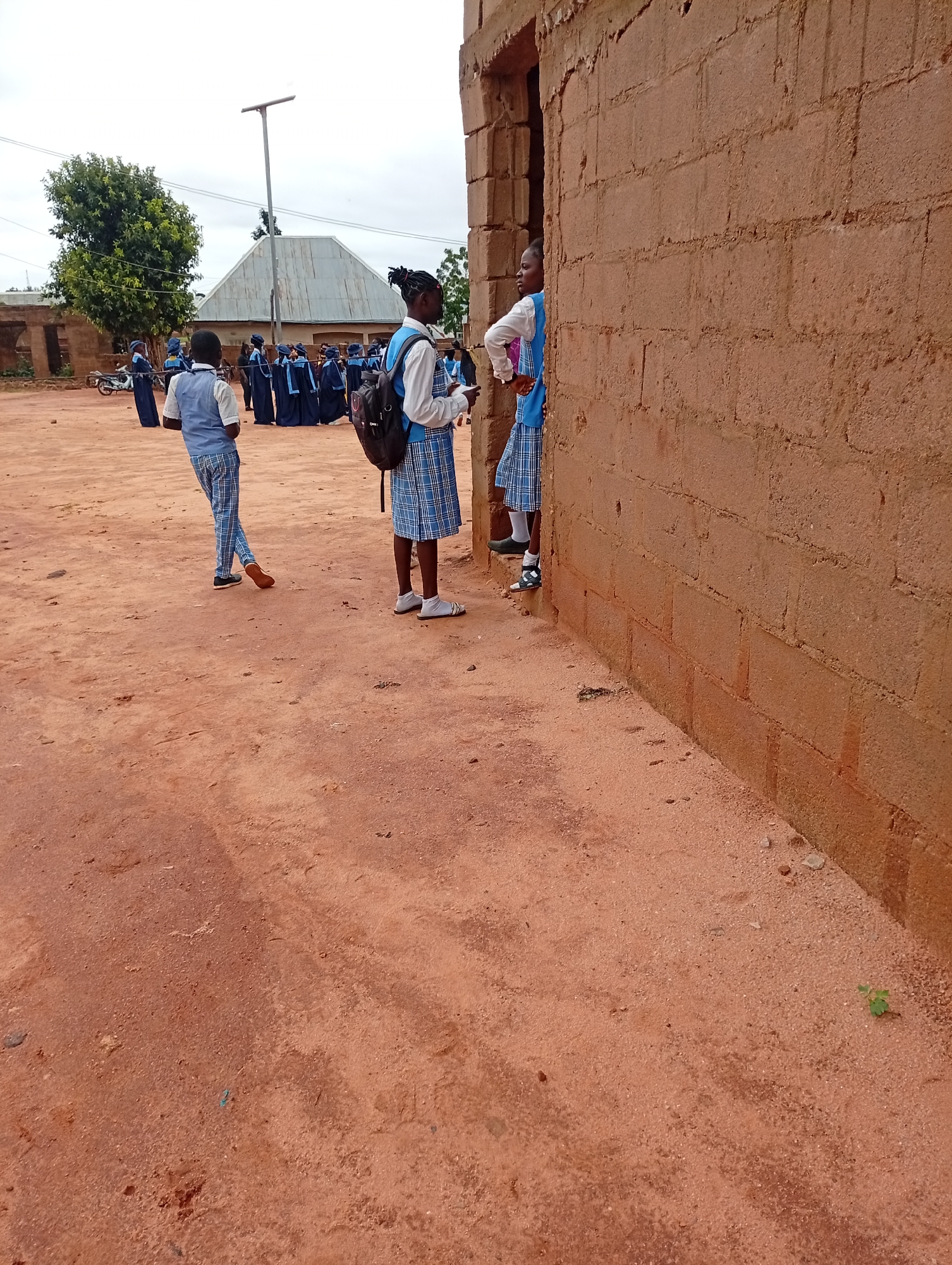 In Tumfure, Nigeria, on an overcast day, a group of students in school uniforms are gathered outside a building constructed of cinder blocks. One student, wearing a light blue vest over a white shirt and plaid pants, stands in the foreground with his left leg extended. Behind him, another student in a similar uniform with a blue plaid skirt stands next to the building's entrance, looking to the left. Further back, a larger group of students in matching blue robes and caps are visible. The ground is covered in red dirt. In the background, a silver-roofed structure, a tree, and a street lamp are present.