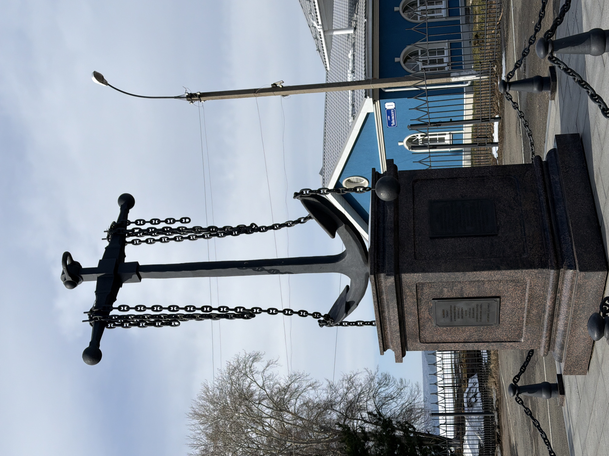 The image depicts the Anchor Monument in Votkinsk, Russia. The monument consists of a large black anchor attached to a dark granite base, which has plaques mounted on it. Heavy chains are draped around the anchor. A blue building with white trim, identified by a street sign as "Tchaikovskogo 1," stands behind the monument. A lamppost rises to the left of the anchor. The ground is paved, and bare trees are visible in the background. A black metal fence runs along the side of the monument, partially obscured by snow. The sky is overcast.
