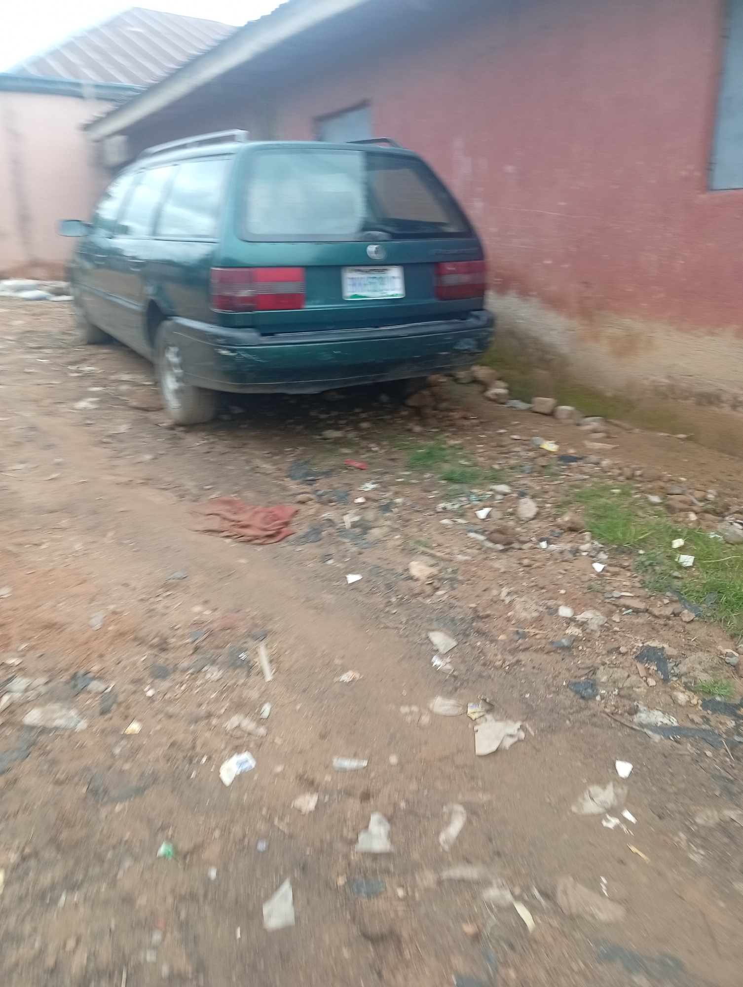 A dark green station wagon is parked on a dirt road in front of a building with a red exterior wall. The car has a roof rack and a license plate that reads "MK539DC". The ground is covered in dirt, rocks, and debris, including some scattered pieces of white paper and plastic. Some patches of green grass are visible. The building appears to be a residential or commercial structure. The sky is overcast. The image seems to be taken in a rural or semi-urban area.