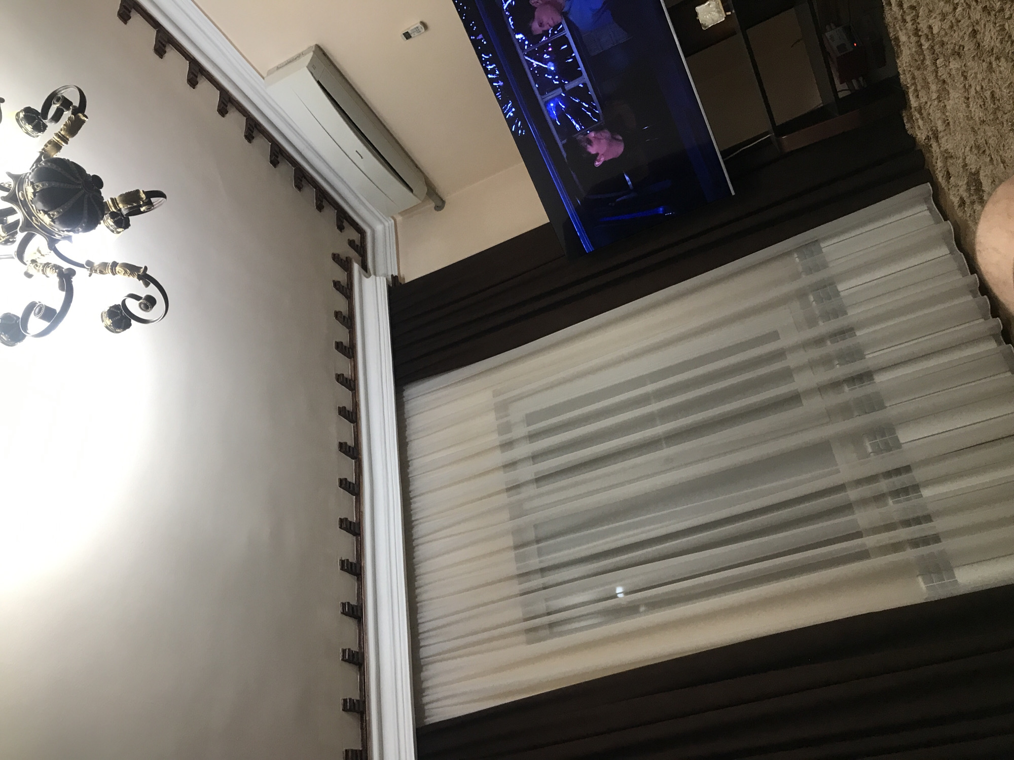 The image shows an interior view of a room. Visible elements include a chandelier on the left side of the image, a wall with decorative molding, a window covered with layered curtains (sheer white fabric over a darker brown fabric), an air conditioning unit mounted near the ceiling, and a television screen showing people on what appears to be a ladder or scaffold with blue lighting. A portion of a carpet and what looks like a person's leg are visible on the right side of the image. There are no clear indications of the image being taken in Yerevan, Armenia.