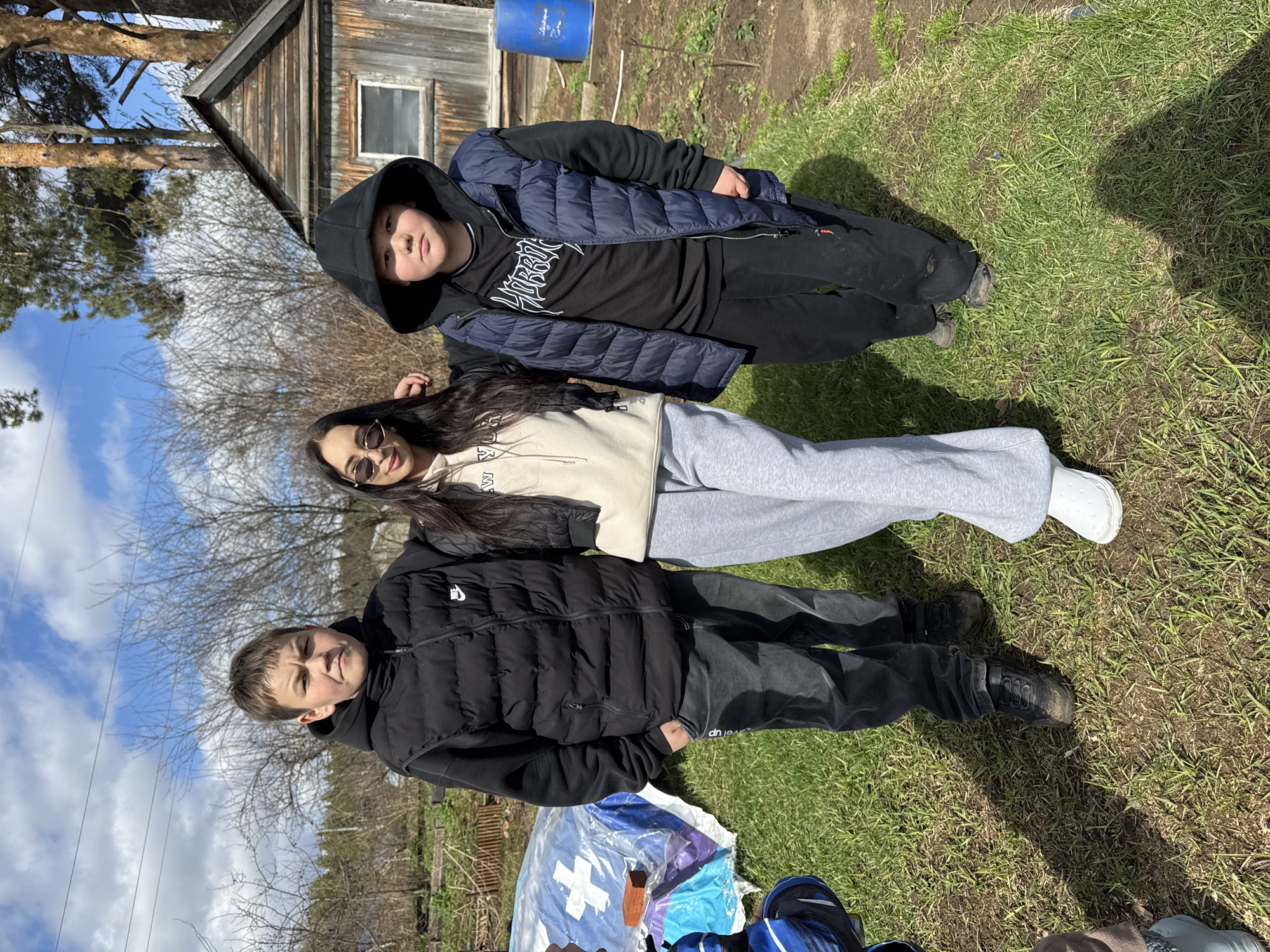 Three people are lying on the ground in Chaikovsky, Russia. The person at the bottom wears a black puffer vest over a dark top and black pants, with black shoes. The person in the middle wears a beige sweater and grey sweatpants, with white shoes. The person on top wears a black hoodie, a blue puffer vest, a black t-shirt, and black pants, with dark shoes. They are on grassy ground near a wooden building with a window. A blue barrel is visible above the building. Trees and a cloudy sky are in the background. Some plastic bags and items are visible near the bottom of the image.
