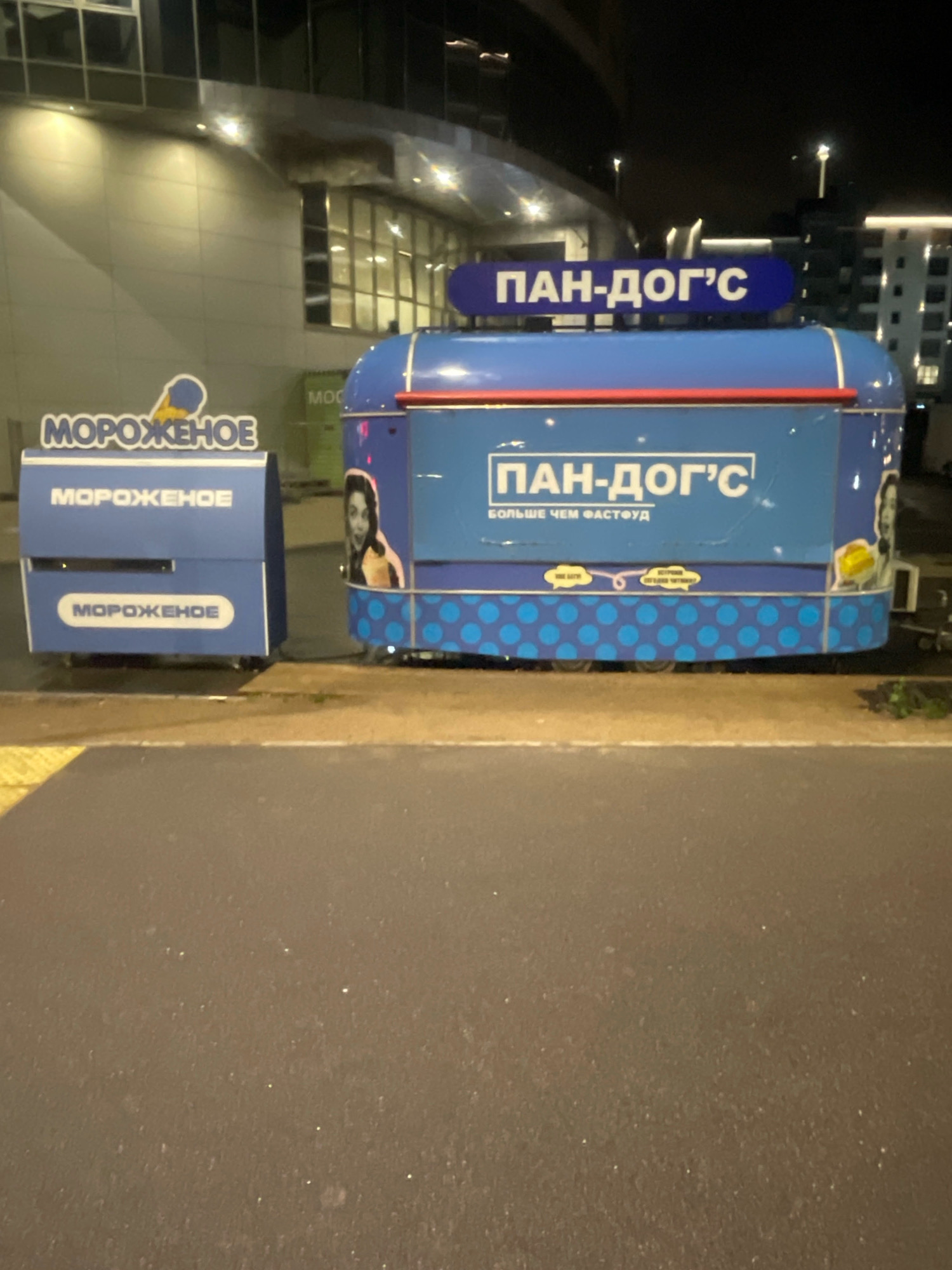 The image depicts an outdoor scene at night, likely in an urban environment. Two food kiosks are visible. The kiosk on the left is blue and white and displays the word "МОРОЖЕНОЕ" (ice cream) multiple times. The kiosk on the right is also blue and features a large blue oval sign with white text reading "ПАН-ДОГ'С" (Pan-Dog's). Below this sign, smaller text reads "БОЛЬШЕ ЧЕМ ФАСТФУД" (More than fast food). A graphic of a person with an ice cream cone is on the side of the right kiosk. Both kiosks appear to be mobile units, suggested by the wheels visible on the right kiosk. In the background, a modern building with large windows and illuminated lights is present, indicating a commercial or office space. The ground in the foreground is a paved surface, possibly a sidewalk or road, with a yellow tactile paving strip. The overall atmosphere is dark, with artificial lighting illuminating the scene. The specific location in Saint Petersburg, Russia cannot be determined from the visual information alone.