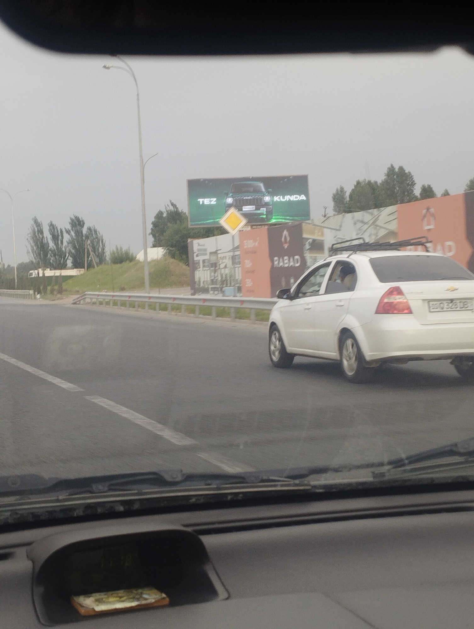 The image depicts a scene from inside a vehicle, looking out onto a road. A white sedan, possibly a Chevrolet, is driving on the right side of the road. The car has a roof rack and a license plate with the inscription "ZQ 328 DB". The interior of the car is visible at the bottom of the frame, showing a dashboard with a small, rectangular object placed in a compartment.
The setting is a highway with a guardrail running alongside the road. In the background, there are trees and a billboard. The billboard displays a car and the words "TEZ KUNDA". Another billboard to the right has the word "RABAD" on it. The weather appears overcast, and the time of day seems to be daytime, judging by the overall lighting.
The image suggests the activity of driving on a highway. The people in the car are not clearly visible, but one person can be seen in the driver's seat of the white sedan. The overall mood of the image is ordinary, capturing a moment during a drive. The location cues, such as the billboard text and the style of the car and road, point to Tashkent, Uzbekistan.