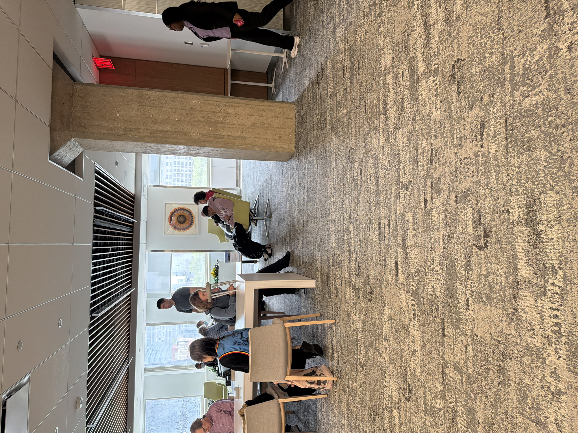The image depicts a modern office or communal space in New York City, USA, as suggested by the urban skyline visible through the windows. Several people are seated at tables, engaged in work or study. The interior features a combination of neutral-toned walls, wooden accents, and a textured carpet, with a large concrete beam serving as a prominent architectural element. The furniture consists of chairs and tables arranged to facilitate group work. Natural light floods the space through large windows. An emergency exit sign is also visible near the top of the frame. A person dressed in a black blazer and pants is walking past the concrete beam on the right. A painting of a floral pattern hangs on the wall.