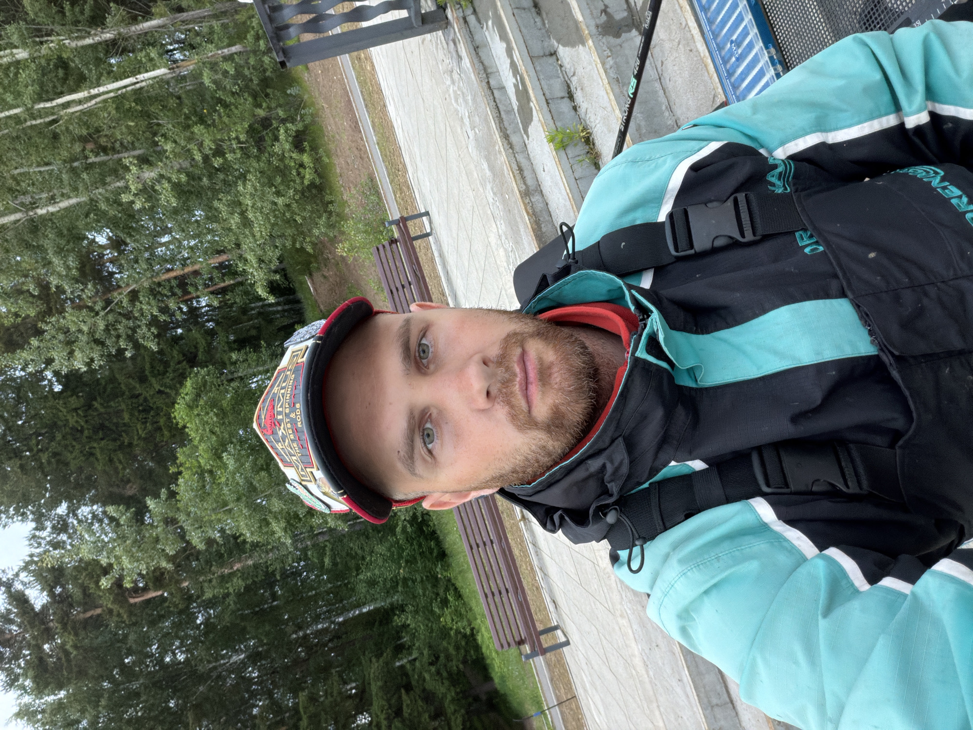 A male individual, approximately 20-35 years old, with light-colored eyes and a brown beard, is centered in a selfie taken outdoors in the city of Chaikovsky, Russia. He wears a multi-colored baseball cap with text including "MAXIM" and "THE BEST SPINNING & RODS" on the front. His attire consists of a teal and black two-piece garment, likely a waterproof jacket and overalls, featuring white vertical stripes on the teal sleeve sections. A red collar is visible beneath the jacket. In the lower right corner, a black fishing rod displaying "STRONG RS 420" is partially visible, alongside a blue object with white stripes, consistent with fishing equipment. The background features a light-colored paved pathway with a brown wooden park bench situated on the left. To the right, concrete steps descend, adjacent to a dark metal railing. The distant background is characterized by a dense forest composed of both coniferous and deciduous trees under a muted sky.