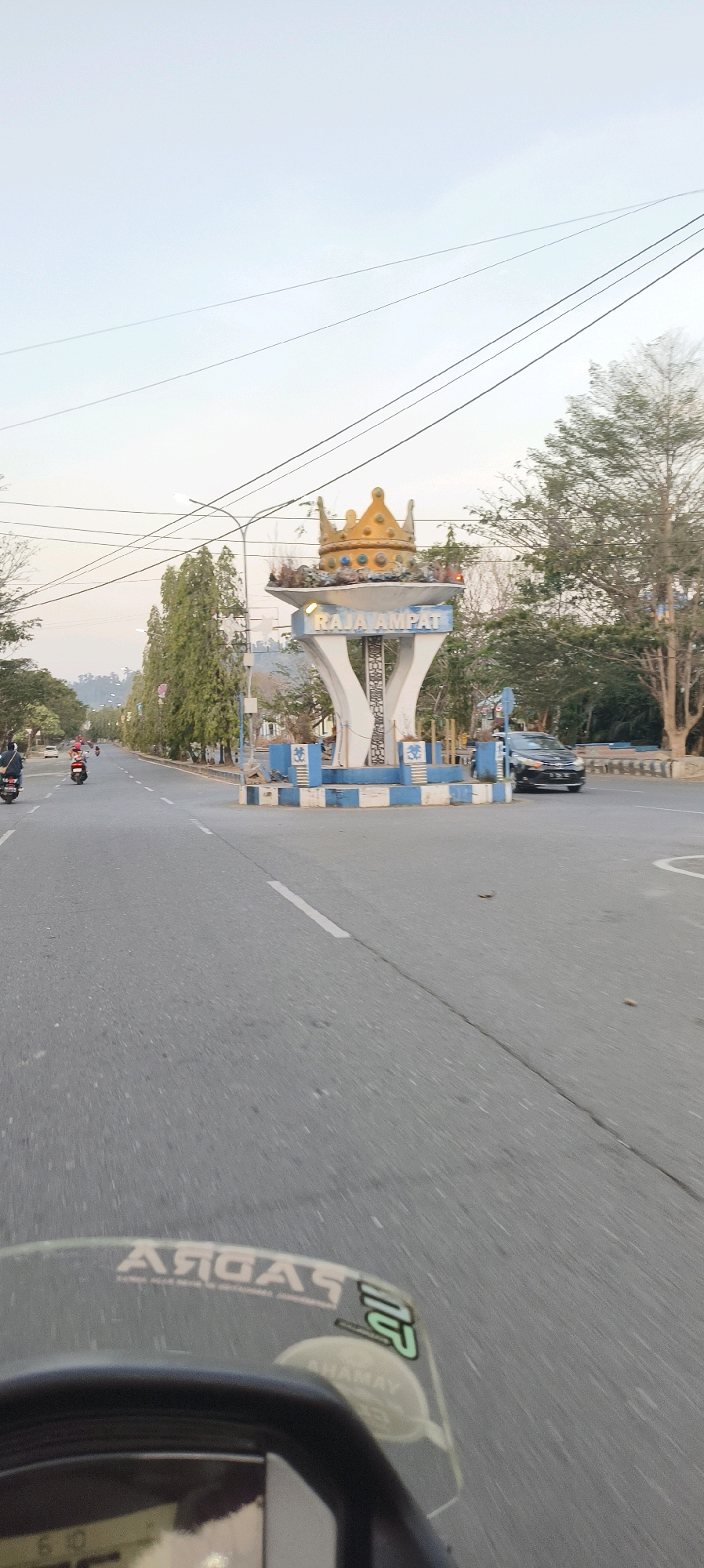 The image shows a street scene in Waisai, Indonesia, featuring a prominent monument topped with a golden crown, which is likely a landmark. The monument has the inscription "RAJA AMPAT" on it, indicating the Raja Ampat Regency. The monument is situated at a road intersection. On the road, several motorcycles and a car are visible, suggesting ongoing traffic. The surrounding environment includes trees lining the street and utility poles with power lines. The sky is overcast, and the overall lighting suggests it is either early morning or late afternoon. The image is taken from the perspective of someone on a moving vehicle, possibly a motorcycle, given the blurry foreground showing the handlebars and windshield of a vehicle with some decals.