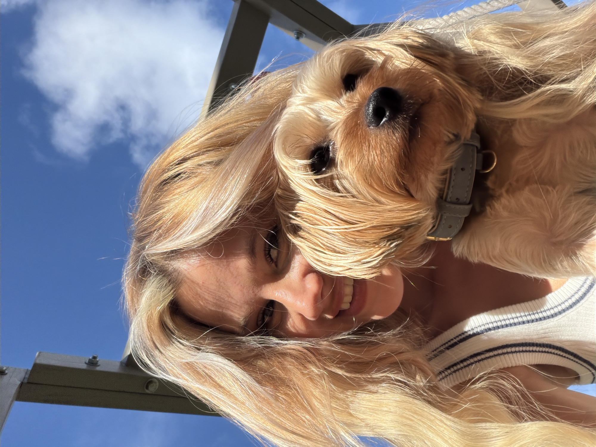 An outdoor, close-up, eye-level, selfie-style photograph shows a young woman with blonde hair smiling and holding a small, light-brown dog with floppy ears. The woman is wearing a white sweater vest with dark blue stripes. The dog is wearing a light grey collar. They are positioned against a bright blue sky with scattered white clouds. A metal structure, possibly part of a playground or outdoor furniture, is visible in the upper left and right portions of the frame. The lighting suggests it is daytime, with sunlight casting shadows on the woman's face. The image does not contain information that would indicate a specific location in Thep Krasatti, Thailand, or any other city.