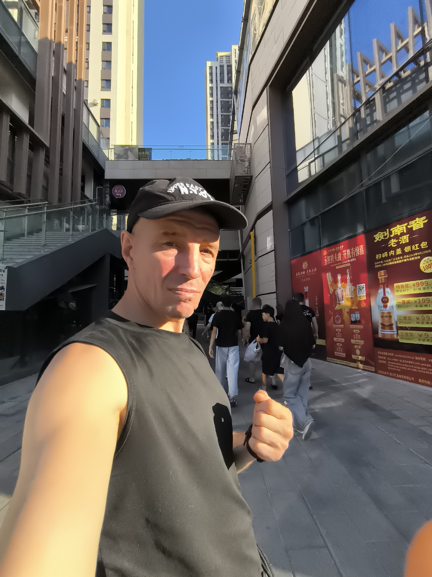 A bald, light-skinned man wearing a black sleeveless shirt and a black baseball cap with "UPPER EAST N.Y.C." embroidered on it is taking a selfie. The man is standing on a paved walkway in front of a modern building with large windows. He is looking directly at the camera with a slight smile. Sunlight is casting a warm glow on his face and arm.
Behind the man, several other people are walking in the same direction. They are dressed in casual clothing. The buildings in the background are tall and modern, with glass facades reflecting the blue sky and surrounding structures. There are some advertisements displayed on a large red banner on the right side of the image, featuring bottles of alcohol and Chinese text. The environment appears to be an urban commercial area.
The image was taken in Ürümqi, China, likely in a commercial district or shopping area, as suggested by the modern architecture, advertising, and presence of pedestrians. The time of day appears to be late afternoon or early evening, given the angle of the sunlight.