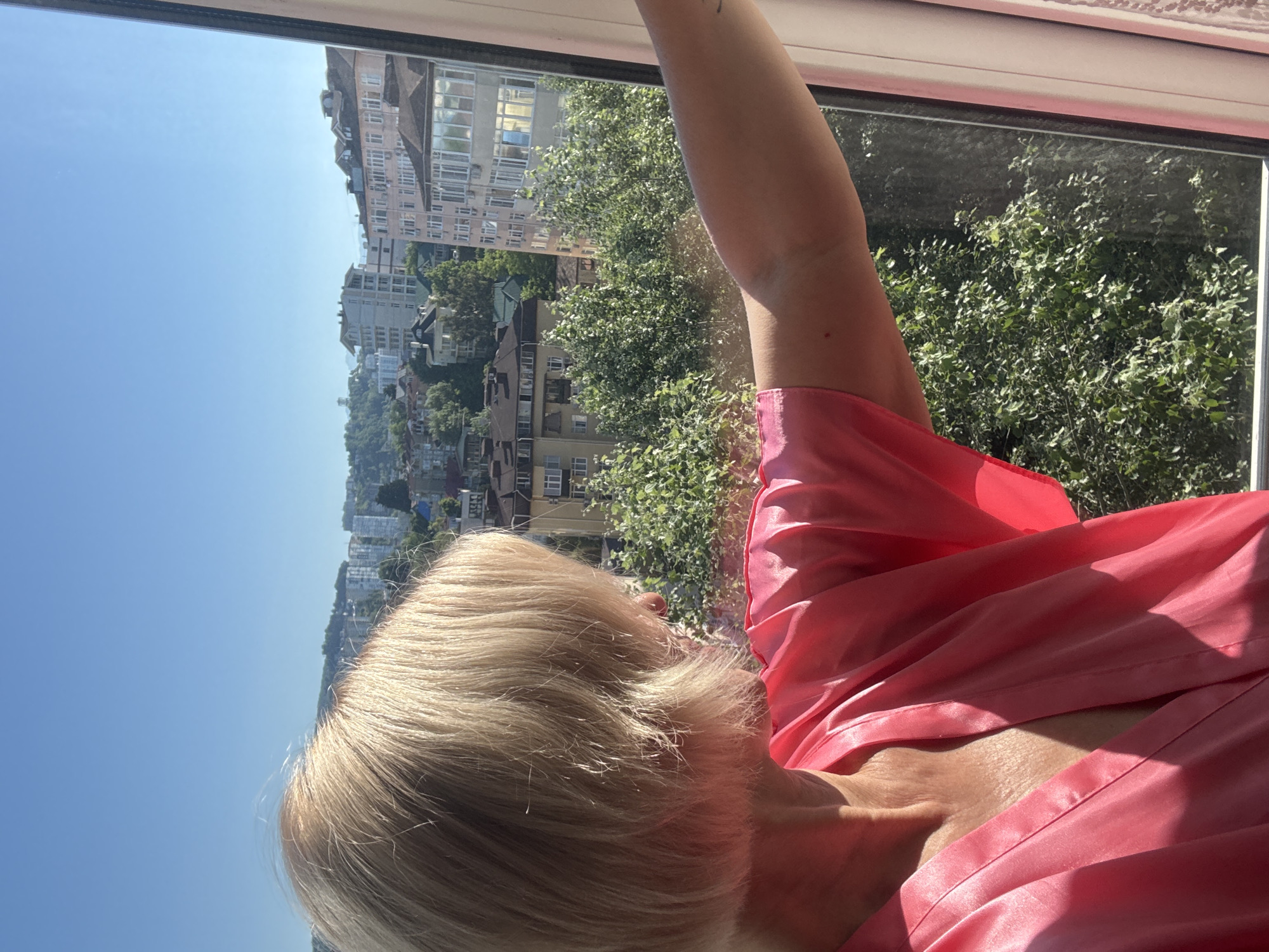 An overhead shot captures a blonde woman in a pink satin robe looking out a window at a cityscape and lush green trees. The woman's face is mostly obscured, but her blonde hair is visible as it is parted to the side. She is wearing a pink robe that drapes around her neck and shoulders. The window offers a view of a city with buildings of varying heights, some with balconies and windows. Trees are abundant throughout the city, creating a green canopy. The sky is a clear blue, indicating a sunny day. The scene appears to be in Sochi, Russia, given the architecture and vegetation typical of the city.