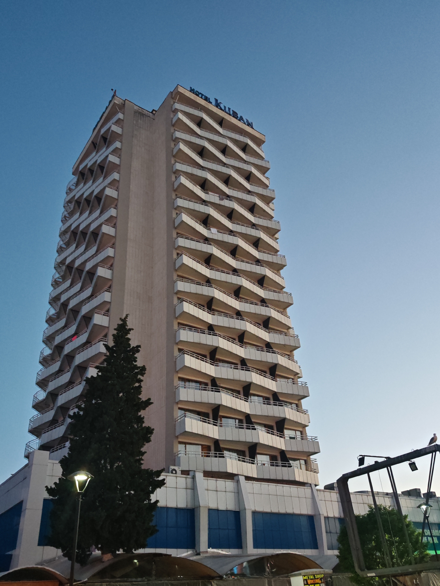 The image shows the Hotel Kuban in Nesebar, Bulgaria, from a low-angle perspective. The tall, rectangular hotel building is the main subject, featuring a concrete central section flanked by numerous floors with zigzagging balconies. The hotel's name is visible at the top in blue letters. A slender, dark green tree stands on the left side of the frame near the base of the hotel. A street lamp is next to the tree. In the lower right, a portion of metal gantry with light fixtures is visible. A bird stands perched on top of the gantry. The sky is light blue and clear.