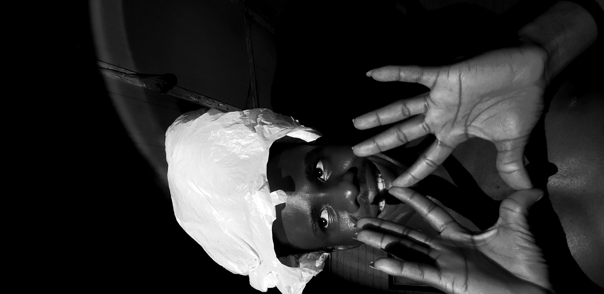 In a dimly lit indoor environment, a Black male, appearing to be in his late teens or early twenties, is captured from the chest up. He is wearing a white plastic bag on his head like a hat. His hands are raised and positioned in front of his face, with fingers interlocked to form a diamond shape between them. He is looking upwards and to his left, with a slight smile on his face, revealing his teeth. The background is dark, with some light casting shadows on the wall and ceiling. The image was taken in Narayi, Nigeria.