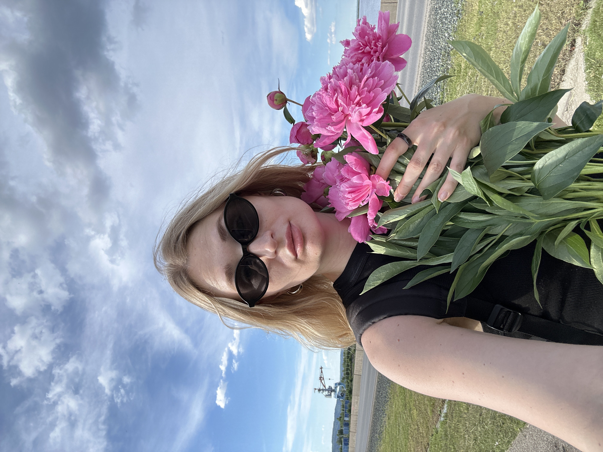 A young woman with blonde hair and dark sunglasses holds a bouquet of pink peonies. She is wearing a black sleeveless top and a black backpack. She is standing outdoors on a sunny day, with a blue sky filled with white clouds above. In the background, there is a body of water and a road with a large industrial crane in the distance. The location appears to be near the Kama River in Chaikovsky, Russia, likely a waterfront area.
