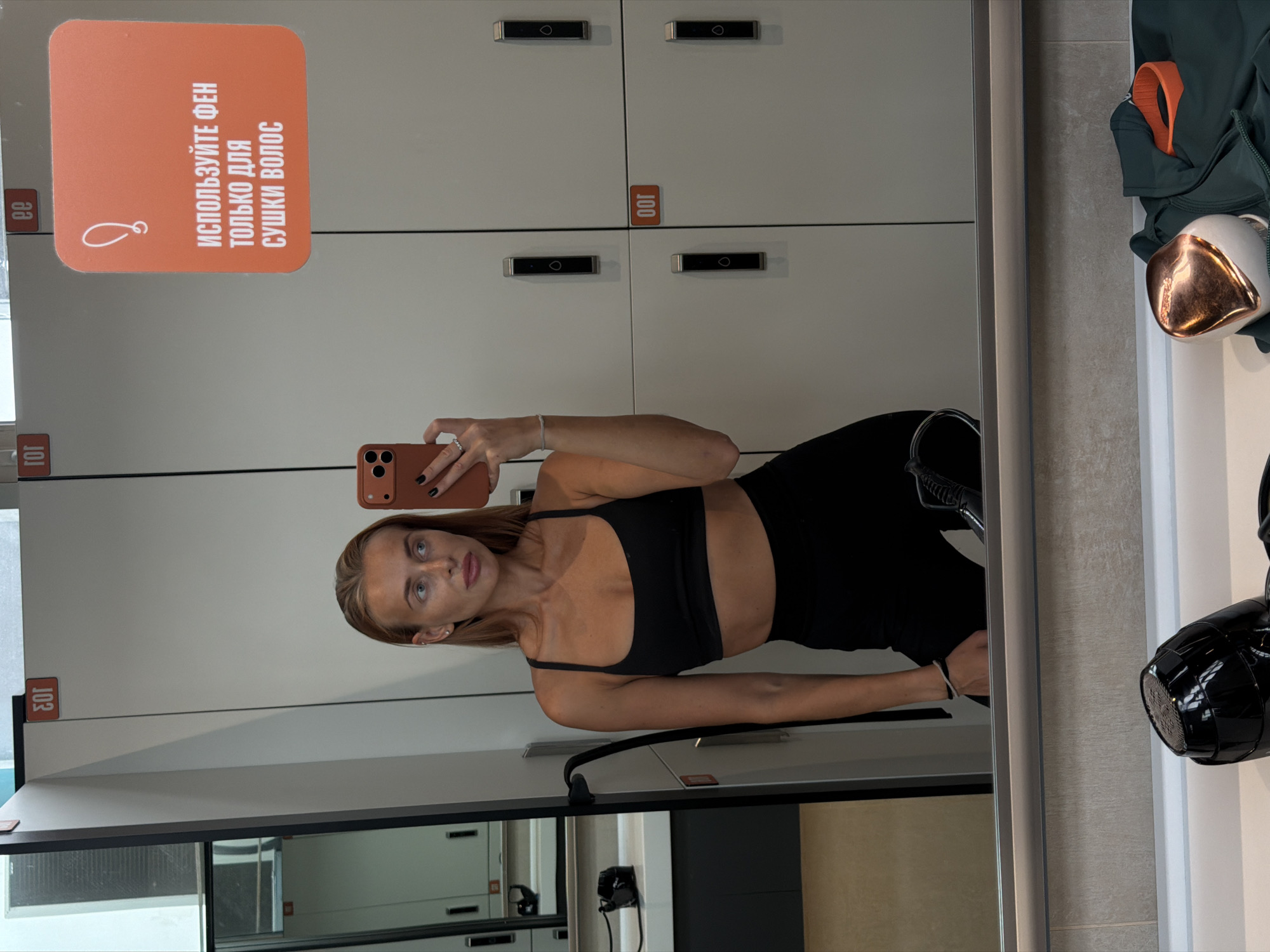 A young woman is taking a selfie in a gym locker room. She is wearing a black sports bra and black leggings. She has blonde hair and is looking at the camera. In the background, there are lockers and a mirror. There is also a hairdryer hanging on the wall. A sign on one of the lockers reads in Russian, "USE THE HAIRDRYER ONLY FOR DRYING HAIR." The woman is smiling slightly. The setting appears to be a modern gym.