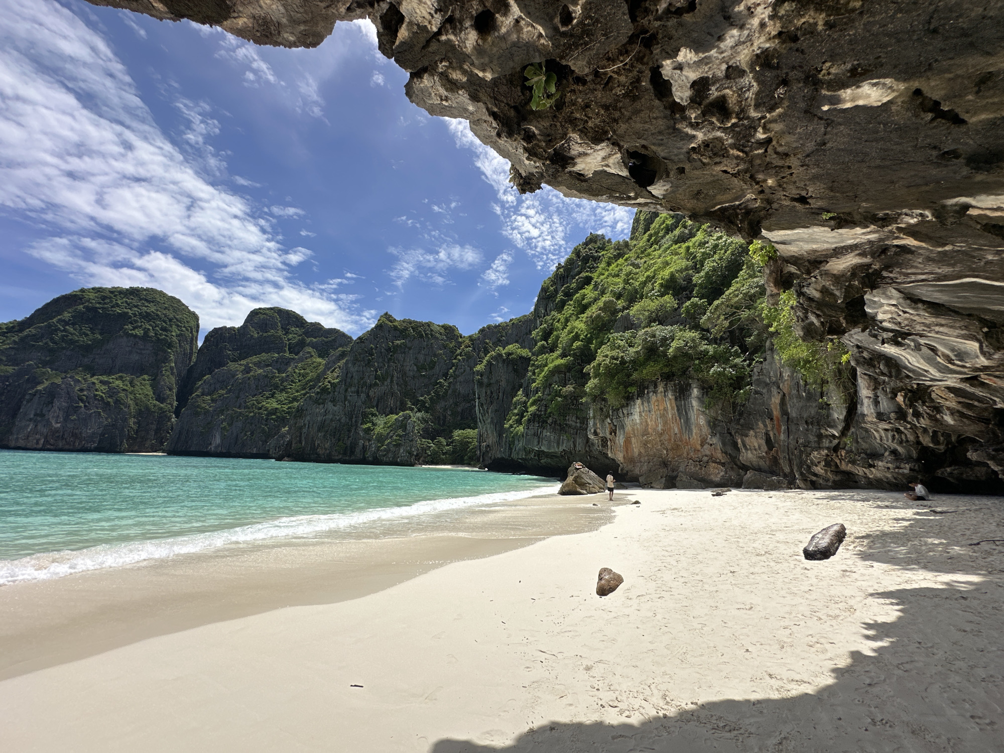 The image shows a beach in Ao Nang, Thailand, with a cave in the foreground. The cave has a rough, rocky texture and is partially covered in vegetation. In the distance, there are lush green cliffs and clear turquoise water. The beach is made of white sand and there are a few people scattered around. One person is walking along the shoreline, and another is sitting near the cave entrance. There is also a piece of driftwood on the sand. The sky is blue with scattered white clouds, and the sun is shining, creating shadows on the beach.