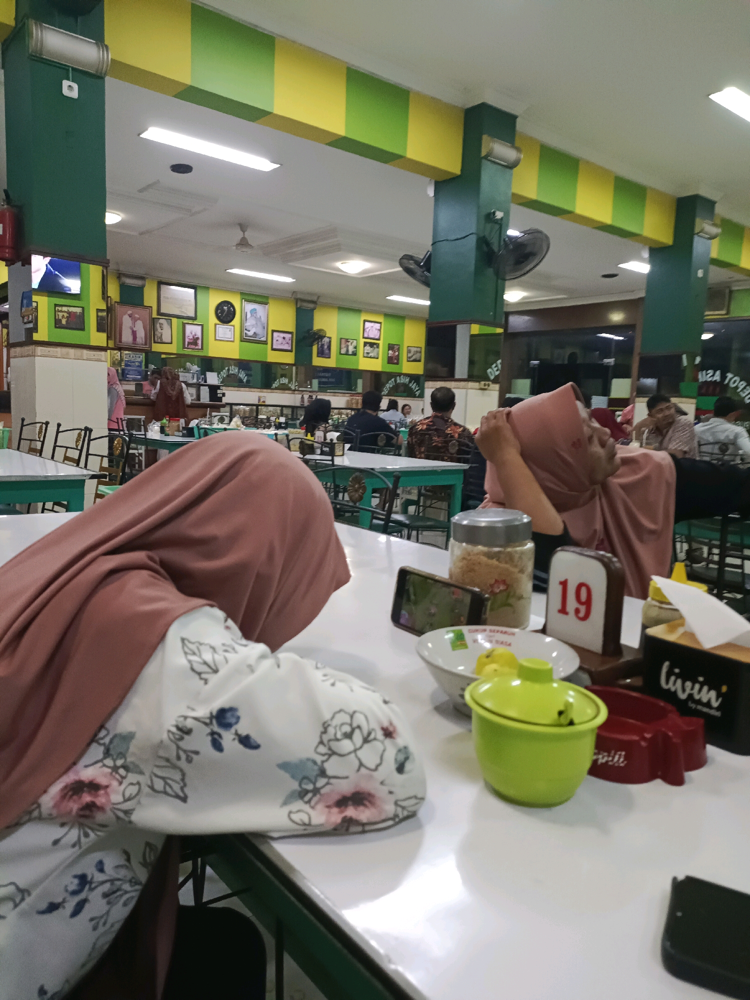 The image depicts an indoor scene at a restaurant, likely in Dapur, Indonesia, identified by signage visible in the background. The restaurant's interior is characterized by a color scheme of green and yellow, with these colors appearing in checkered patterns on beams and walls. In the foreground, a person wearing a pink hijab and a white floral-patterned shirt is seated at a white table. Their face is not fully visible as they are turned away from the camera. On the table, various items are arranged, including a glass jar filled with a light-colored granular substance, a smartphone displaying an image, a white bowl with yellow items, a bright green container with a lid, a red condiment holder, and a black tissue box with the word "livin'" printed on it. A table number "19" is also prominently displayed. In the mid-ground, other patrons are visible seated at tables. Some are engaged in conversation or eating. In the background, shelves filled with items and more framed pictures adorn the walls. Two ceiling fans are visible, and fluorescent lighting illuminates the space. A fire extinguisher is mounted on a green pillar on the left side of the image. The overall atmosphere appears to be casual and busy, typical of a local eatery.