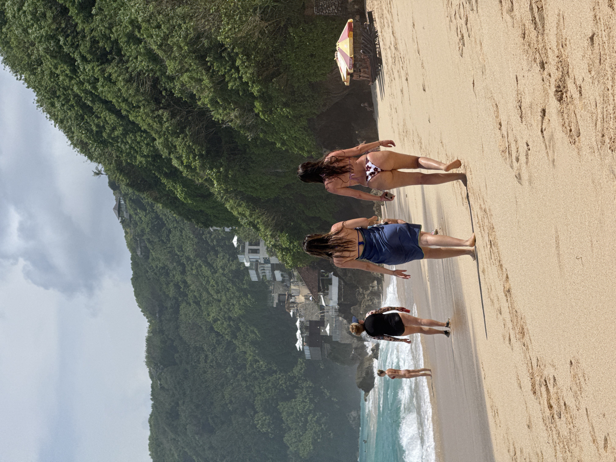 The image captures a vibrant beach scene in Kuta Selatan, Indonesia, looking towards lush, green cliffs. The primary subjects are several people enjoying the beach, mostly viewed from behind.
In the foreground, three women are walking on the light brown sand, heading towards the left side of the frame. The woman closest to the camera, on the right, wears a floral-patterned bikini bottom and a light-colored top, with long dark hair. To her left and slightly ahead, another woman wears a blue dress or cover-up over swimwear, also with long dark hair. Further left and slightly behind them, a third woman is dressed in a black swimsuit or shorts and top, displaying tattoos on her arm/shoulder, and appears to have lighter hair. Near the waterline on the far left, another person in light swimwear is visible.
The sandy beach, marked with footprints and subtle shadows, stretches across the midground. On the right, close to the cliff, a yellow and red striped beach umbrella is partially visible. The background is dominated by towering, verdant cliffs covered in dense tropical foliage. Several white and light-colored buildings are nestled into the cliffside, suggesting coastal residences or resorts. The sky is partly cloudy with patches of grey. On the far left, the ocean meets the shore with white-capped waves.
The scene depicts a daytime setting, likely late morning or early afternoon, with bright natural light. There is no visible text in the image.
