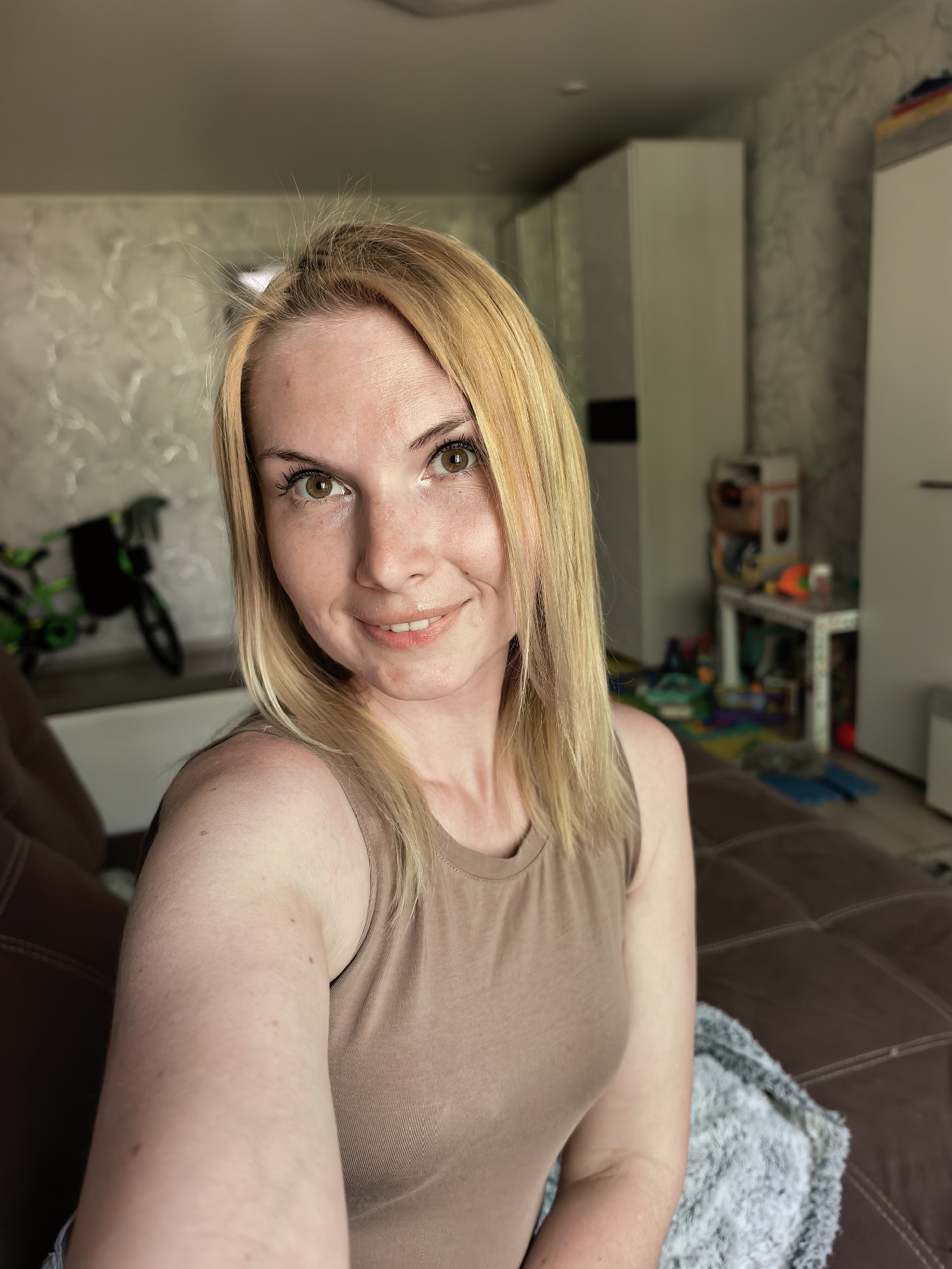 A fair-skinned woman with blonde hair and green eyes is sitting on a brown leather sofa. She is wearing a brown tank top. Behind her is a room with light-colored wallpaper and a white wardrobe. In the background, there is a child's bicycle and various toys scattered around a table. The location is Sterlitamak, Russia.