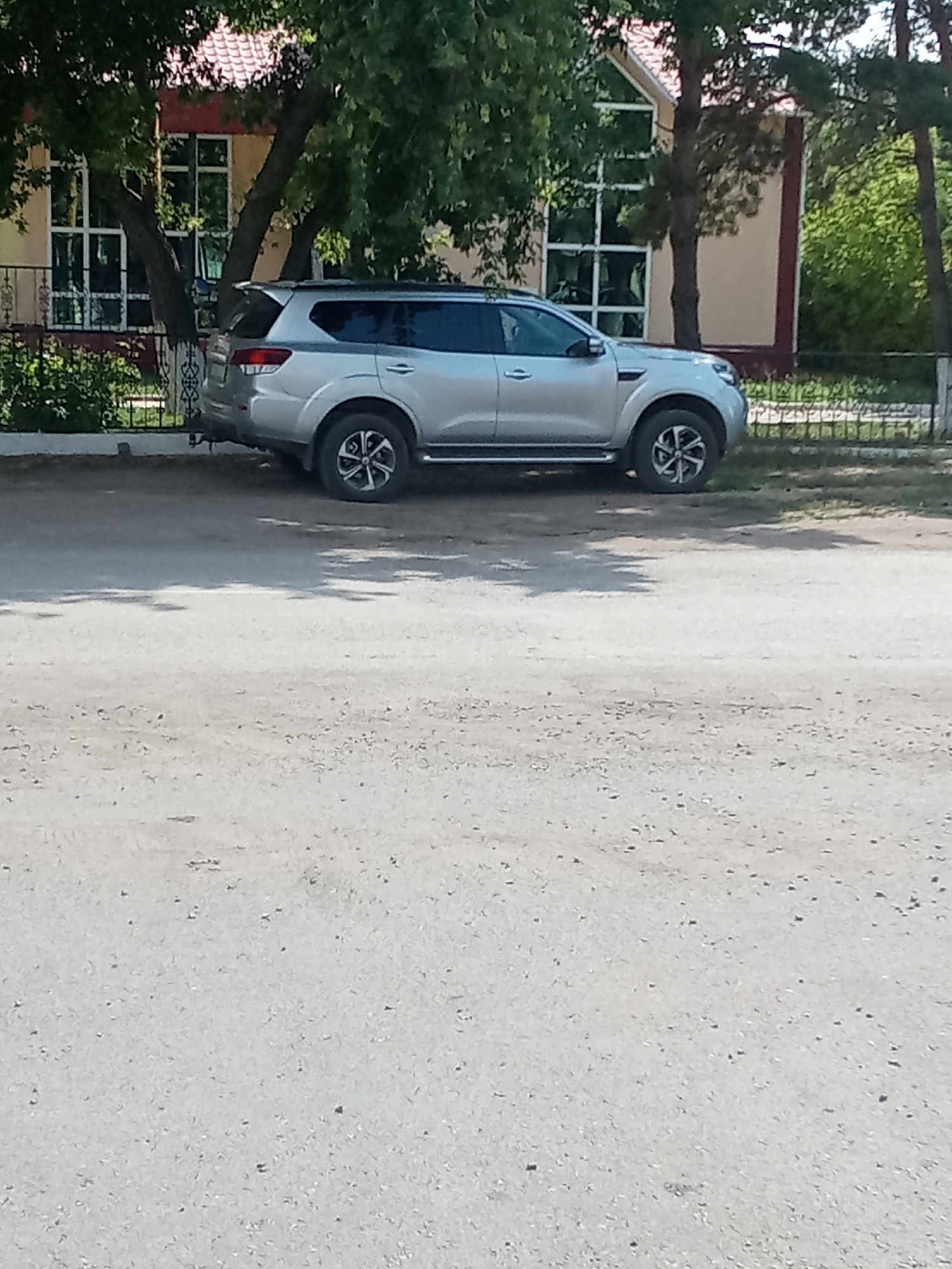 The image captures a silver SUV parked on a gravel lot in front of a building with large windows and a red-tiled roof. Trees with green foliage surround the building and cast shadows on the ground. A black metal fence separates the lot from the building. The SUV is positioned in the center of the frame, showing its side profile. The ground is covered in a mix of sand and small stones, indicating an outdoor, possibly rural or semi-urban, environment. The sky is not visible in the frame.