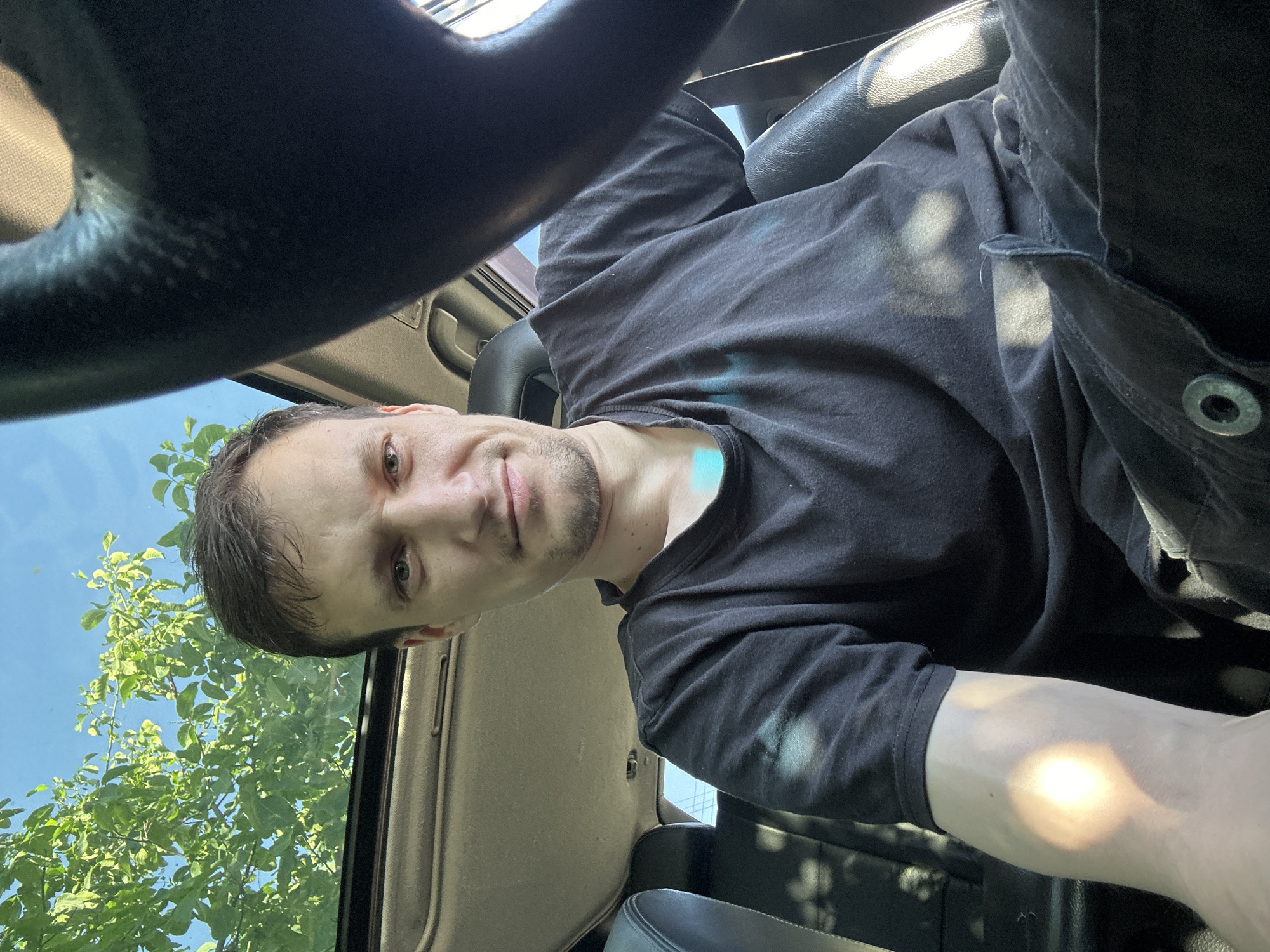 The image is a close-up, selfie-style shot of a man sitting in the driver's seat of a vehicle. He is looking directly at the camera with a neutral expression. The man is wearing a black t-shirt and dark pants. The interior of the car is visible, including the steering wheel and part of the dashboard. Sunlight streams through the open sunroof, illuminating the man's face and arm, and casting dappled shadows from tree leaves onto the interior. The background shows a bright blue sky and green foliage, suggesting the vehicle is outdoors, likely in a park or residential area. The presence of the steering wheel and driver's seat indicates the man is in the driver's seat of a car. The image does not provide any specific visual cues to pinpoint the location within Shatava, Ukraine.