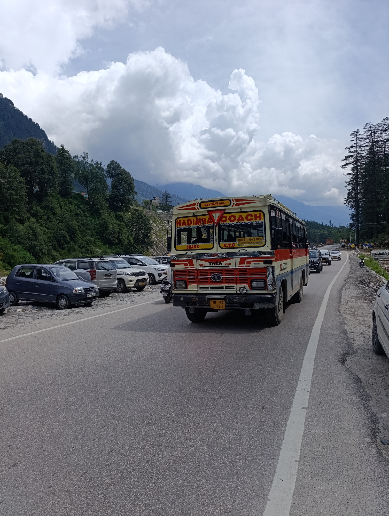 In Chichoga, India, a vintage "HADIMBA COACH" bus with a red, white, and yellow exterior is seen driving on a paved road under a cloudy sky. The bus has "K.T.C" branding and a license plate reading "HP 34 B 1125". Parked on the side of the road are several cars, including a blue hatchback and a white SUV. The background features a lush green hillside dotted with trees, and mountains shrouded in mist under a dramatic cloud formation. The road is a two-lane asphalt surface with a white dividing line.