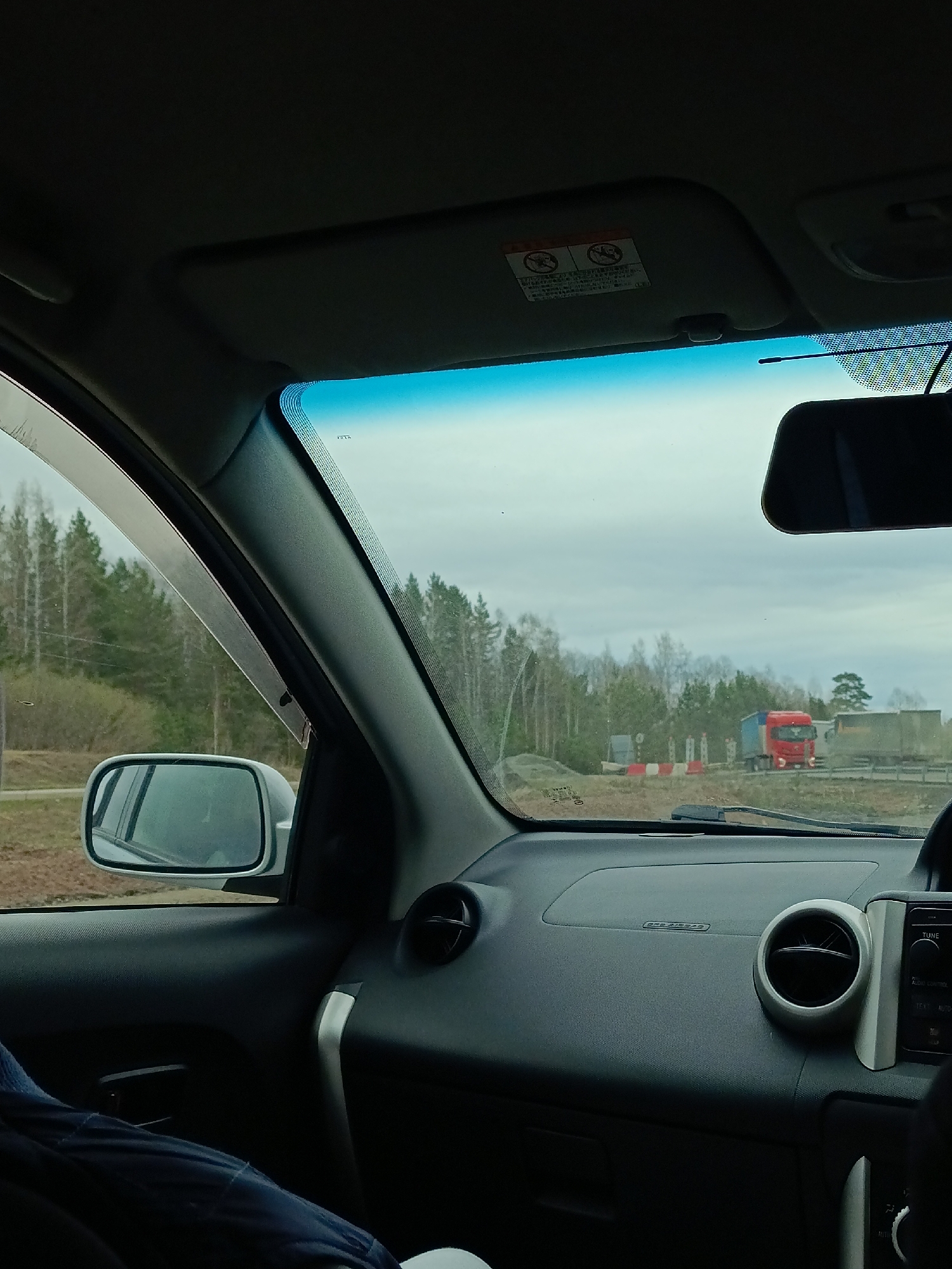 The image shows the view from inside a car, likely a right-hand drive vehicle, looking out onto a road with a weigh station or border checkpoint. Several trucks are visible in the distance, one with a red cab. The environment appears rural with trees lining the road. The overcast sky suggests a cloudy day.
Given the potential location of Malaya Kuskunka, Russia and border crossing point, the checkpoint in the distance, based on available geographical information is possibly linked to the M52 motorway that connects Russia with Mongolia, if so this would imply a significant amount of commercial and industrial truck transportation traffic passing this point.
