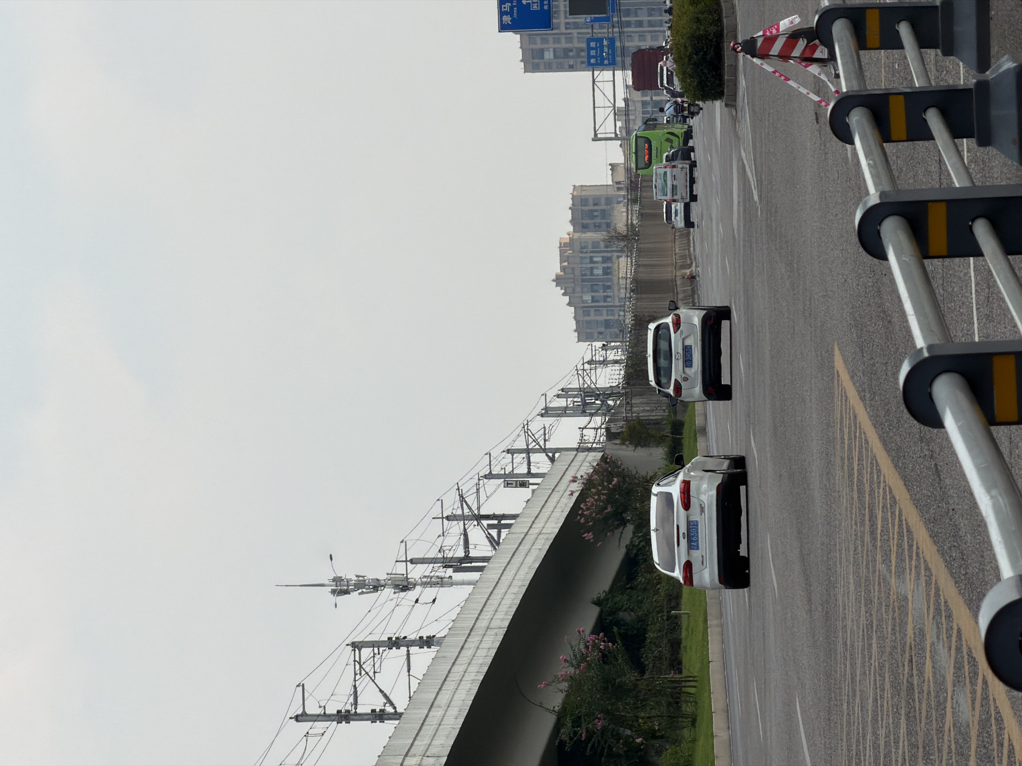 This image captures a view of a multi-lane road in Jiangjin, China. The road is flanked by a raised concrete structure with overhead electrical wires, likely part of an elevated train line or power infrastructure. On the right side of the road, a blue traffic sign with Chinese characters is visible, indicating directions. Several vehicles are on the road, including white cars, a green bus, and a red truck. In the foreground, a metal barrier with black and yellow accents separates the traffic lanes from what appears to be a pedestrian area. The sky is overcast, suggesting a cloudy day. The overall scene depicts an urban transportation artery with active vehicular traffic.