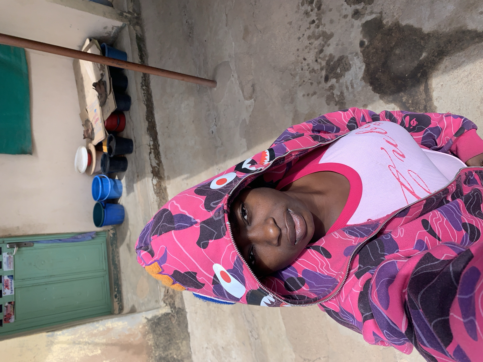 A person with dark skin and dark hair wearing a pink and purple hooded jacket and a pink shirt is photographed from above. The person is facing the camera and smiling slightly. The background consists of a concrete floor with dark stains, a brown metal bar attached to a wall, a green door with items hanging on it, and a stack of buckets and a cardboard box in a corner. The location is Rafin Par, Nigeria, as specified in the prompt.