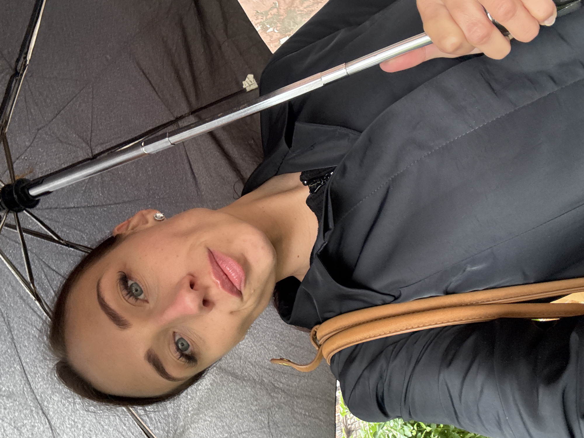 A woman, with fair skin, blue eyes, and dark brown hair pulled back, is taking a selfie outdoors, holding a black umbrella over her head. She is wearing a black jacket and a tan strap from her bag is visible over her left shoulder. Her makeup includes mascara, eyeliner, and pink lipstick. She is wearing a small, diamond stud earring in her left ear. The background appears to be an urban environment with some greenery and a brick building. It is raining, as evidenced by the umbrella and the droplets on its surface. The specific location in Saint Petersburg, Russia is not discernible from the image.
