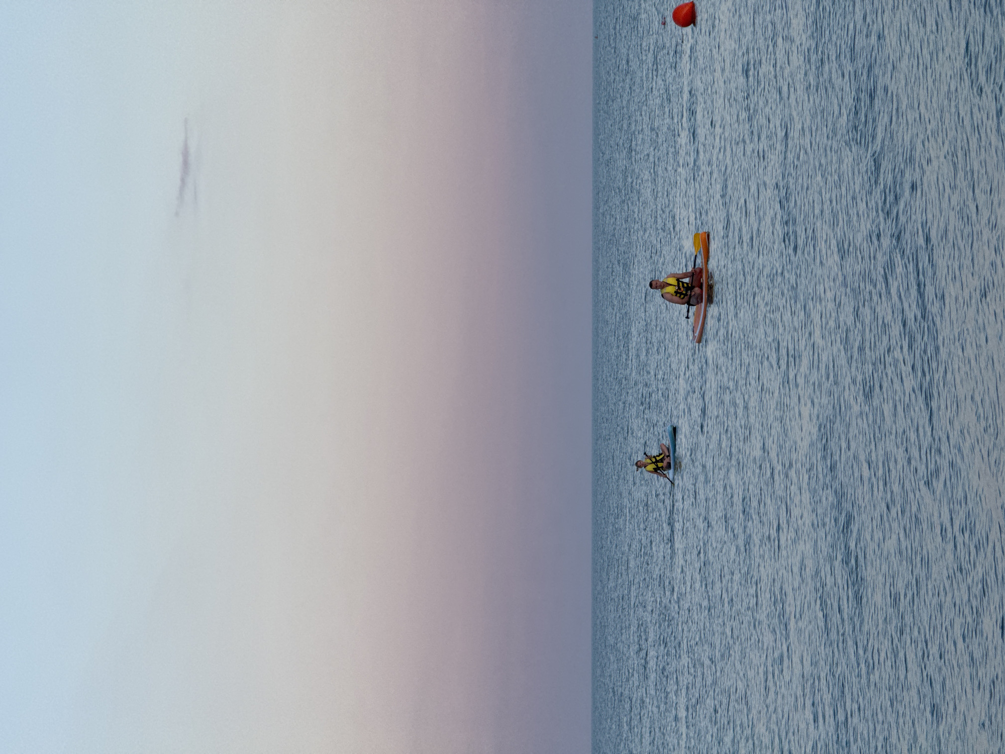 Two people are paddleboarding on the sea. The person on the left is on a blue paddleboard and is wearing a yellow life vest. The person on the right is on an orange paddleboard and is also wearing a yellow life vest. Both individuals are facing left. There is a red buoy to the right of the image. The sky is a gradient of pale blue to pink, indicating it is either sunrise or sunset. The sea is calm and blue. This scene is likely on the coast of Sochi, Russia, given the calm water and clear sky.