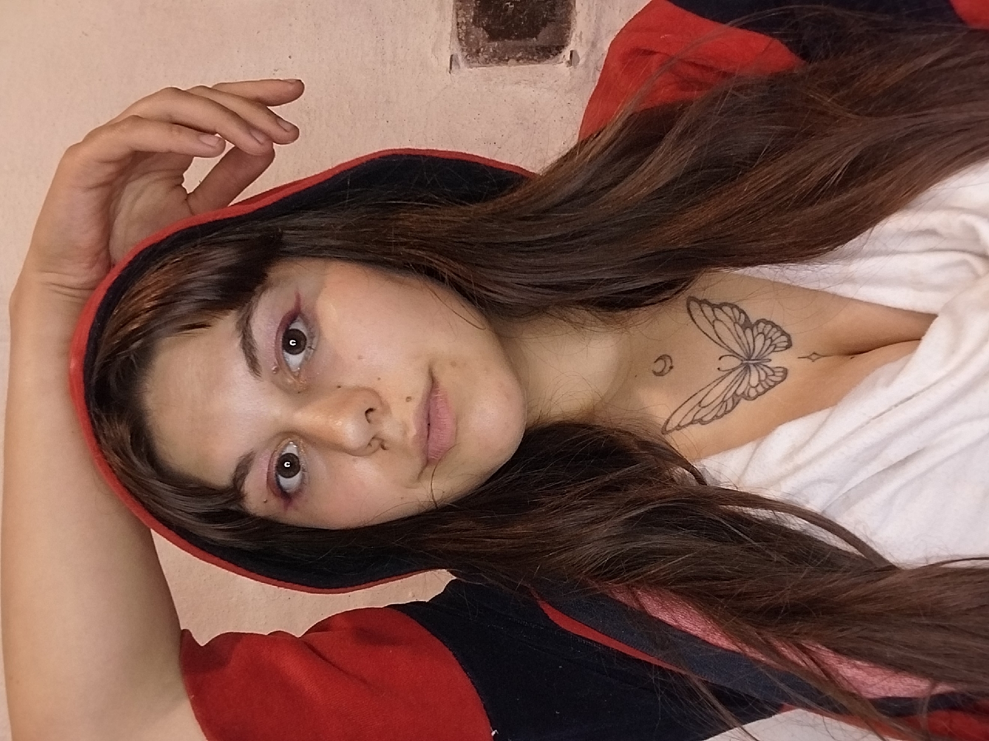 A young woman with dark brown hair and bangs is captured in a close-up shot, her left arm raised and resting on her head. She is wearing a red and black hooded jacket over a white top, which reveals a butterfly tattoo on her chest and a crescent moon tattoo above it. Her makeup includes pink eyeshadow and eyeliner. The background appears to be a textured wall. The image is taken indoors, in an unknown location.