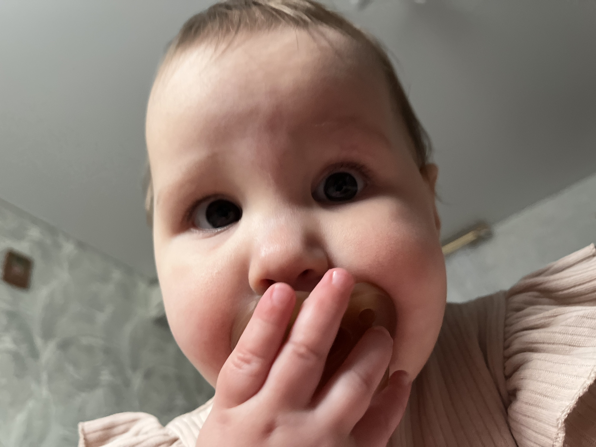 A close-up shot depicts a baby with fair skin, brown hair, and wide dark eyes. The baby has a pacifier in their mouth and is holding it with their hand. The baby is wearing a light pink ribbed top. The background shows a room with textured walls and a decorative framed object on the wall. The setting is not explicitly identified as Novopokrovskaya, Russia, but is a domestic interior.
