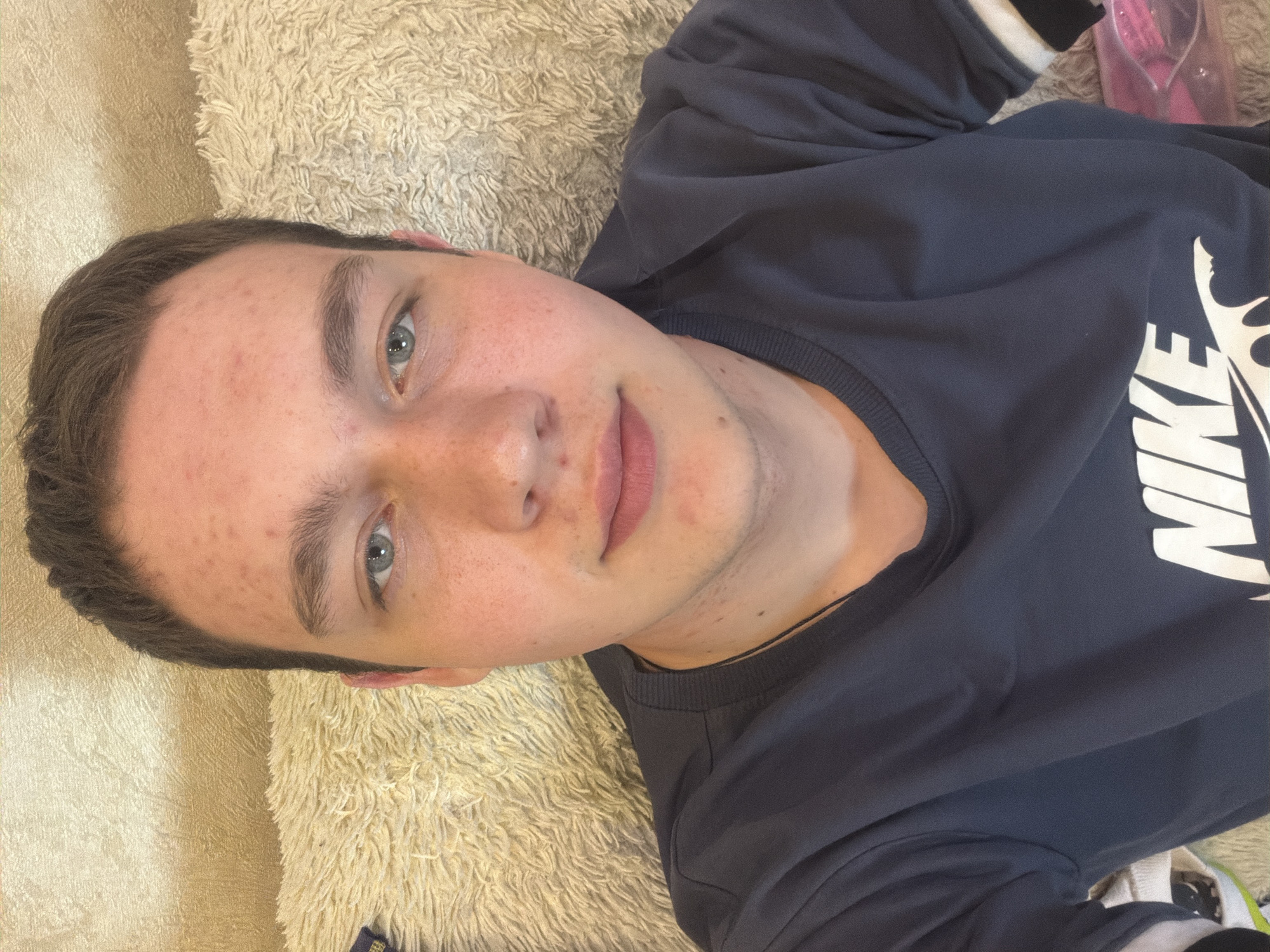 A young man with fair skin and short brown hair is lying down, his head resting on a plush, light-colored surface. He has blue eyes and appears to be looking at the camera. He is wearing a dark blue Nike sweatshirt. The setting appears to be indoors. The location in the city of Severodvinsk, Russia is not evident from the image.