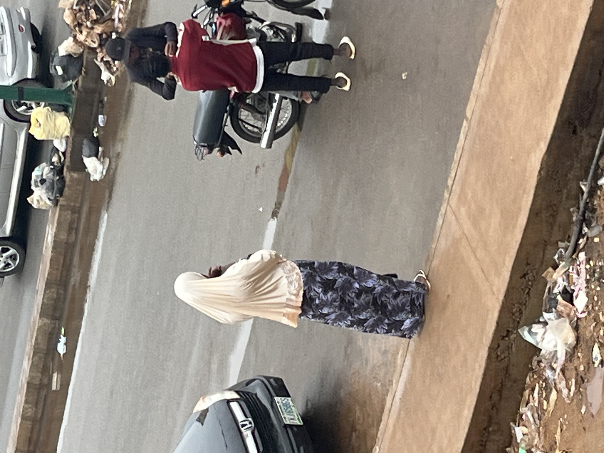The image captures a street scene in Jos, Nigeria, under overcast conditions. A woman wearing a light-colored hijab and a long, dark skirt with a floral pattern stands facing away from the camera, appearing to be in conversation with someone off-frame. To her right, two men are interacting near a motorcycle. One man, wearing a dark jacket and a baseball cap, is holding a mobile phone. The other man, in a maroon jacket and black trousers, stands with his back to the camera. The ground is wet, suggesting recent rain, and scattered debris, including plastic bags and other refuse, is visible along the roadside and on a concrete ledge. A dark-colored car is parked to the left of the frame.