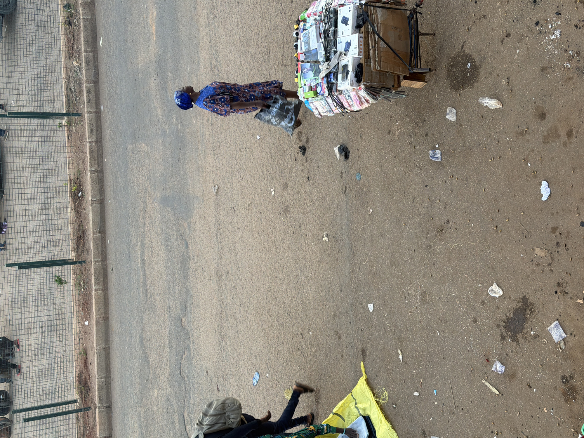 In Bukuru, Nigeria, a woman wearing a blue headscarf and a blue and white patterned dress stands beside a mobile cart laden with various electronic accessories, including phone cases and chargers. She holds a black plastic bag. The cart appears to be a makeshift stall, constructed with cardboard boxes and a metal frame, displaying a colorful assortment of goods.
The scene is set on a paved road, with a wire fence running along the left side of the image. The ground is mostly dirt and asphalt, scattered with small pieces of debris and trash, suggesting a public space, possibly a street market or roadside vendor area. Sunlight indicates it is daytime.
Further down the road, another individual is partially visible, lying on a yellow sheet spread on the ground. This person is dressed in dark clothing and has a light-colored head covering. The overall impression is of a bustling, possibly commercial, outdoor environment in Nigeria.