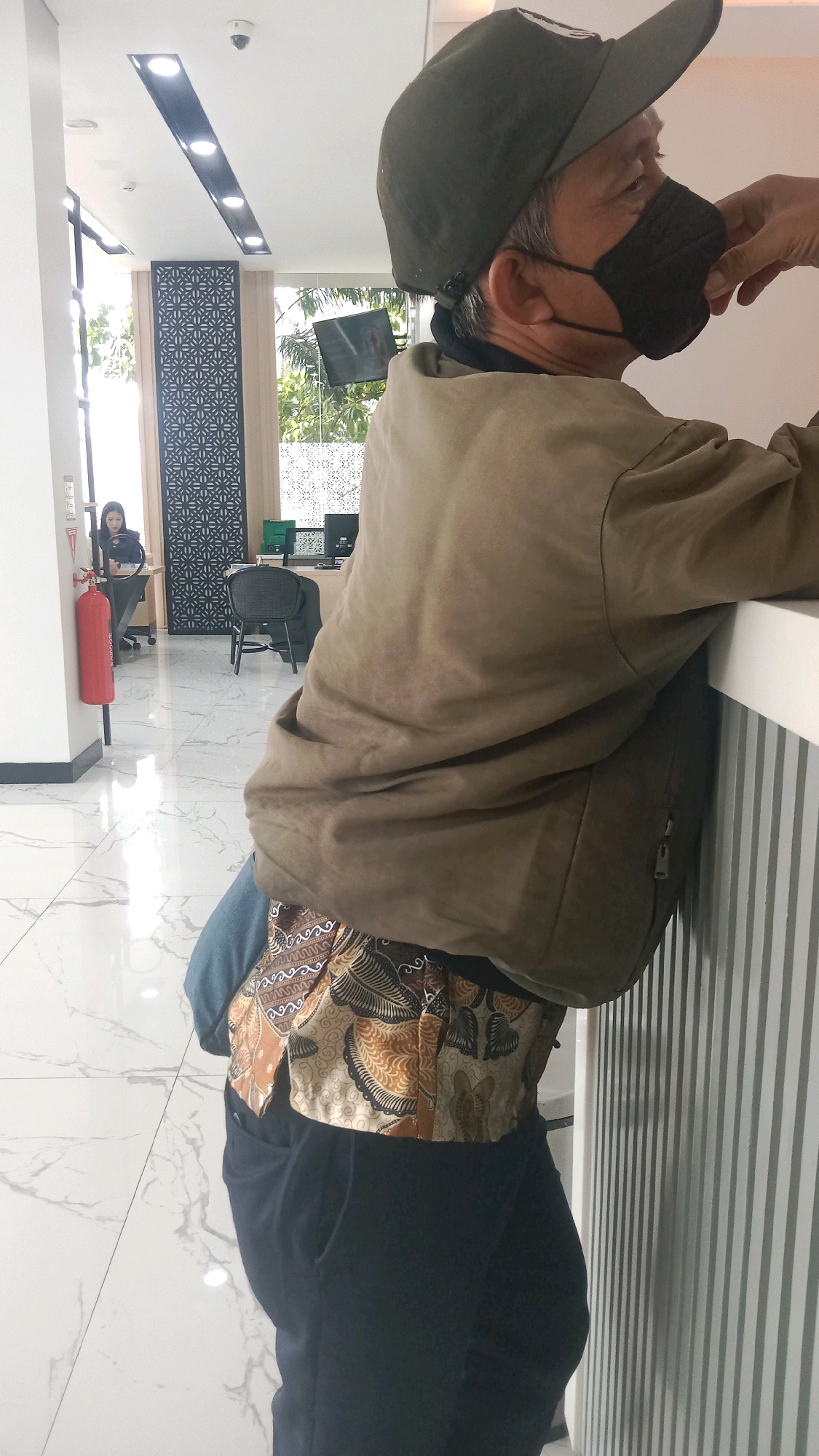 In a well-lit interior space, likely an office or a bank in Bekasi, Indonesia, a person wearing a dark green cap, a black face mask, and a tan jacket is leaning against a white striped wall. Their left hand is touching the face mask. Beneath the jacket, a patterned fabric with brown and beige designs is visible, paired with dark pants. In the background, a woman in a headscarf is seated at a desk, working on a computer. A red fire extinguisher is mounted on a white pillar to the left of the frame. The floor is polished white tile with a marble pattern. The environment appears modern and clean.