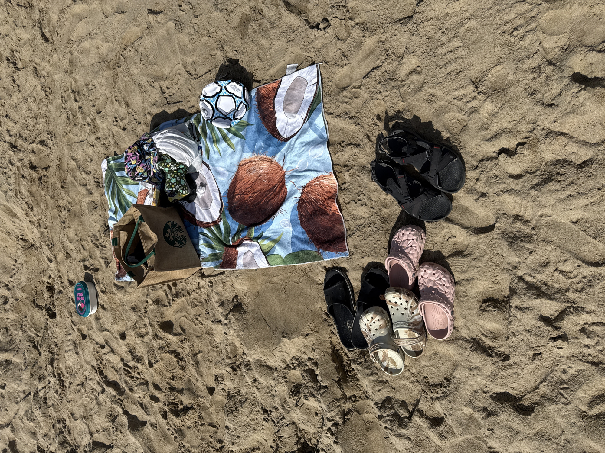 A beach blanket with a coconut print is spread on the sand. On the blanket are a brown canvas tote bag with a Starbucks logo, a stack of folded clothes, and a small soccer ball. Several pairs of footwear are scattered on the sand around the blanket, including black slide sandals, patterned clogs, pink clogs, and black hiking sandals. A small blue and white container is also visible on the sand near the tote bag. The scene appears to be a casual gathering or a break from activities on a sunny beach in Nha Trang, Vietnam, based on the tropical theme of the blanket and the general beach environment.