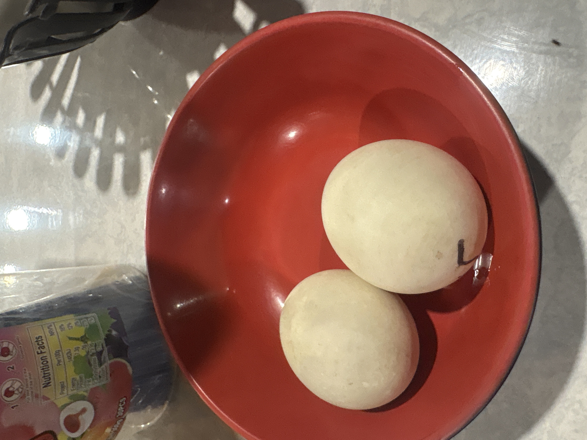 Two light-colored eggs are positioned within a bright red bowl, which is resting on a grey, speckled surface. The bowl is the primary focus of the image. In the background, a plastic package with visible "Nutrition Facts" and ingredient information is partially visible on the left. Shadows from an unseen object, possibly a chair, are cast across the surface behind the bowl. The setting appears to be an indoor location, possibly a dining table. There is no indication of the city of Carmen, Philippines, in the image.