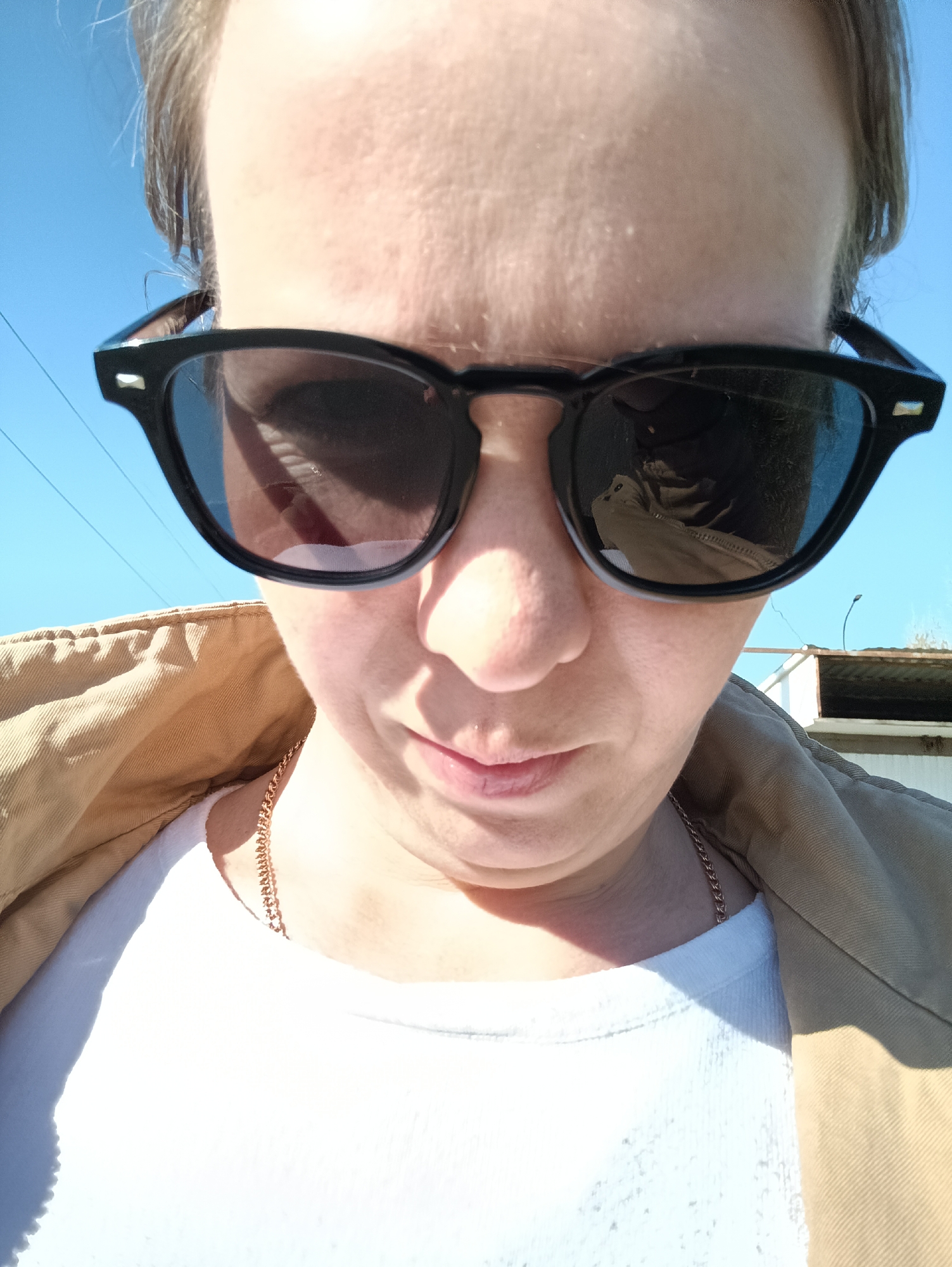 A close-up, low-angle shot of a person with fair skin wearing sunglasses with a black frame. The sunglasses reflect the surrounding environment, including a light-colored jacket, a white t-shirt, and the blue sky. A thin gold chain is visible around the person's neck. The background includes a portion of a white building or structure. Based on the information provided, the location is Chaikovsky, Russia, but there are not enough details in the image to determine anything more specific.