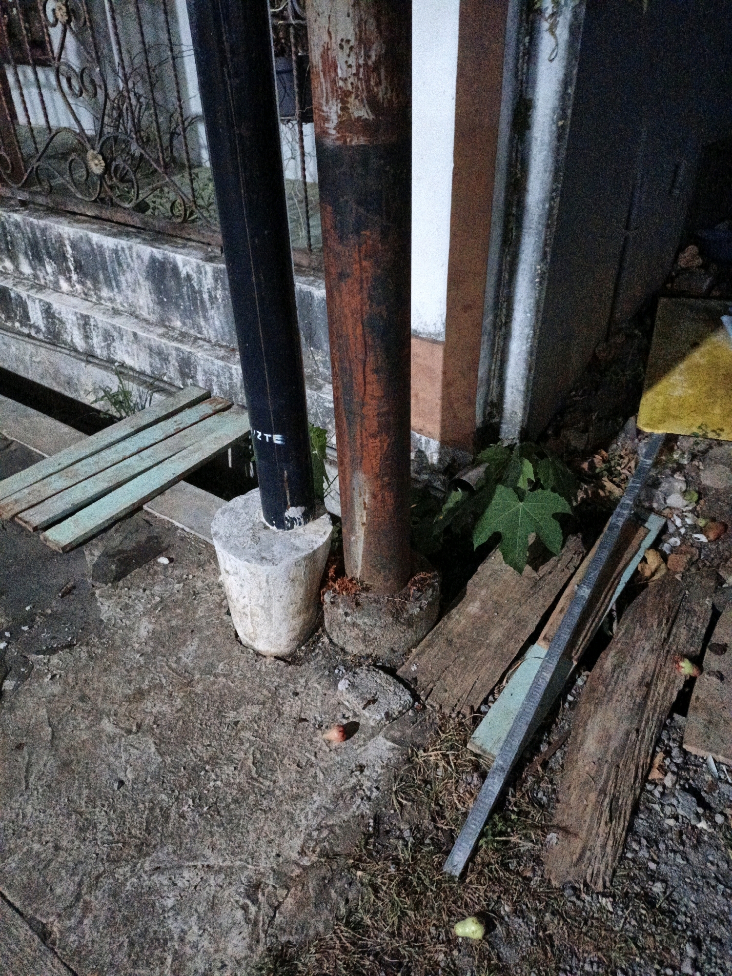 The image captures a close-up, ground-level view of an outdoor scene, likely a sidewalk or alleyway in Cikal, Indonesia, at night. Dominating the center are two vertical posts: one a dark, cylindrical pole, and the other a rusty, reddish-brown metal pipe. The dark pole is partially embedded in a rough, cylindrical concrete base. To the left, several weathered, pale green wooden planks are arranged parallel to each other, spanning a gap. In the background, a weathered, textured concrete wall or structure with a metal railing and some vegetation is visible. To the right of the posts, there is an accumulation of debris, including dirt, dry leaves, and pieces of wood. A small, green leafy plant is growing near the base of the posts. The ground surface is a rough, gray concrete or asphalt with scattered gravel and small stones. A few small, round objects, possibly fallen fruit, are present on the ground. The lighting suggests artificial light sources, creating shadows and highlighting the textures of the materials. No people are visible in the frame.