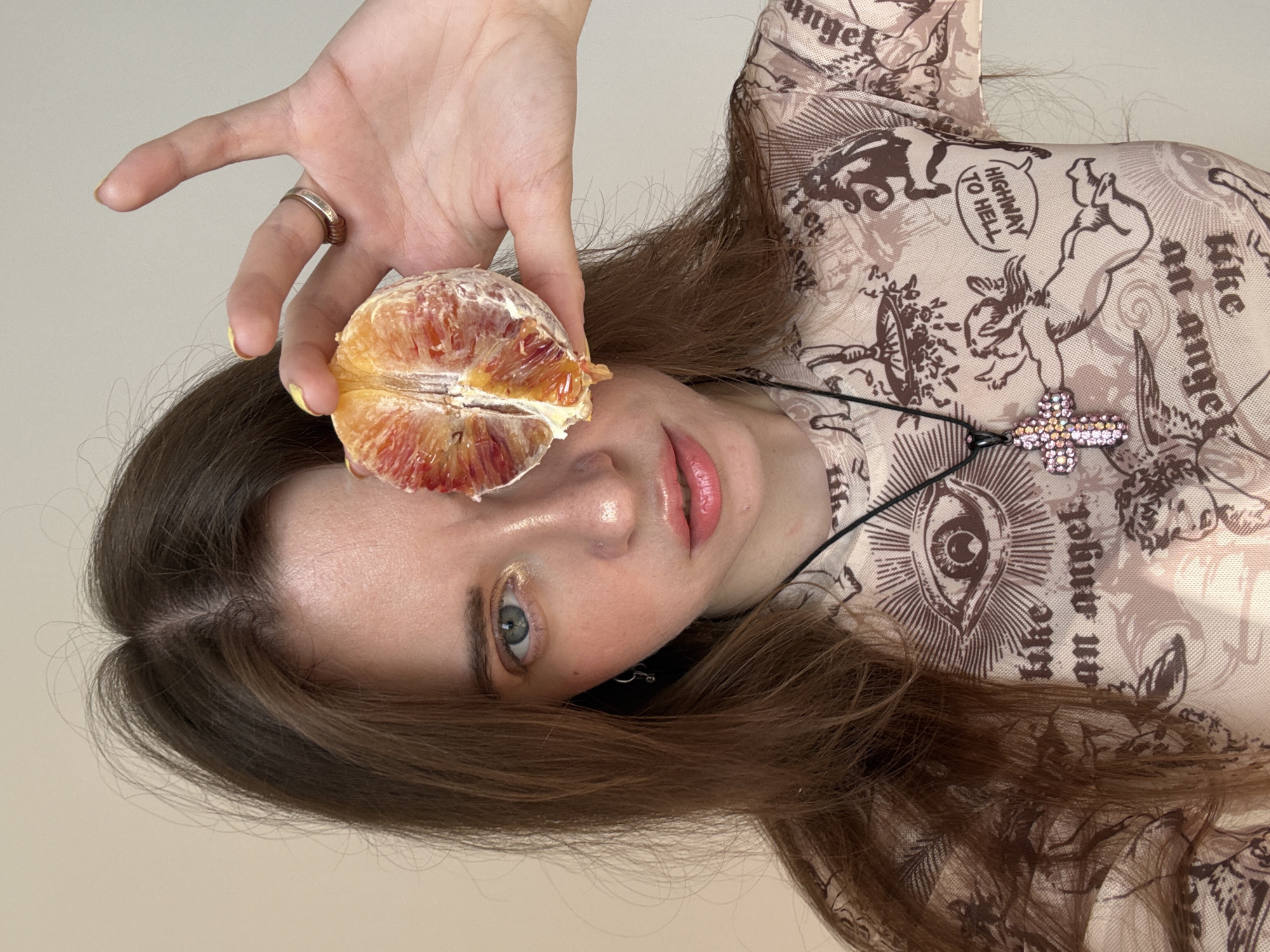 A woman with fair skin and brown hair is holding a halved blood orange in front of her face. She is wearing a sheer, patterned top with a cross pendant necklace. The background is a plain, light-colored wall. The location is not discernible from the provided information.